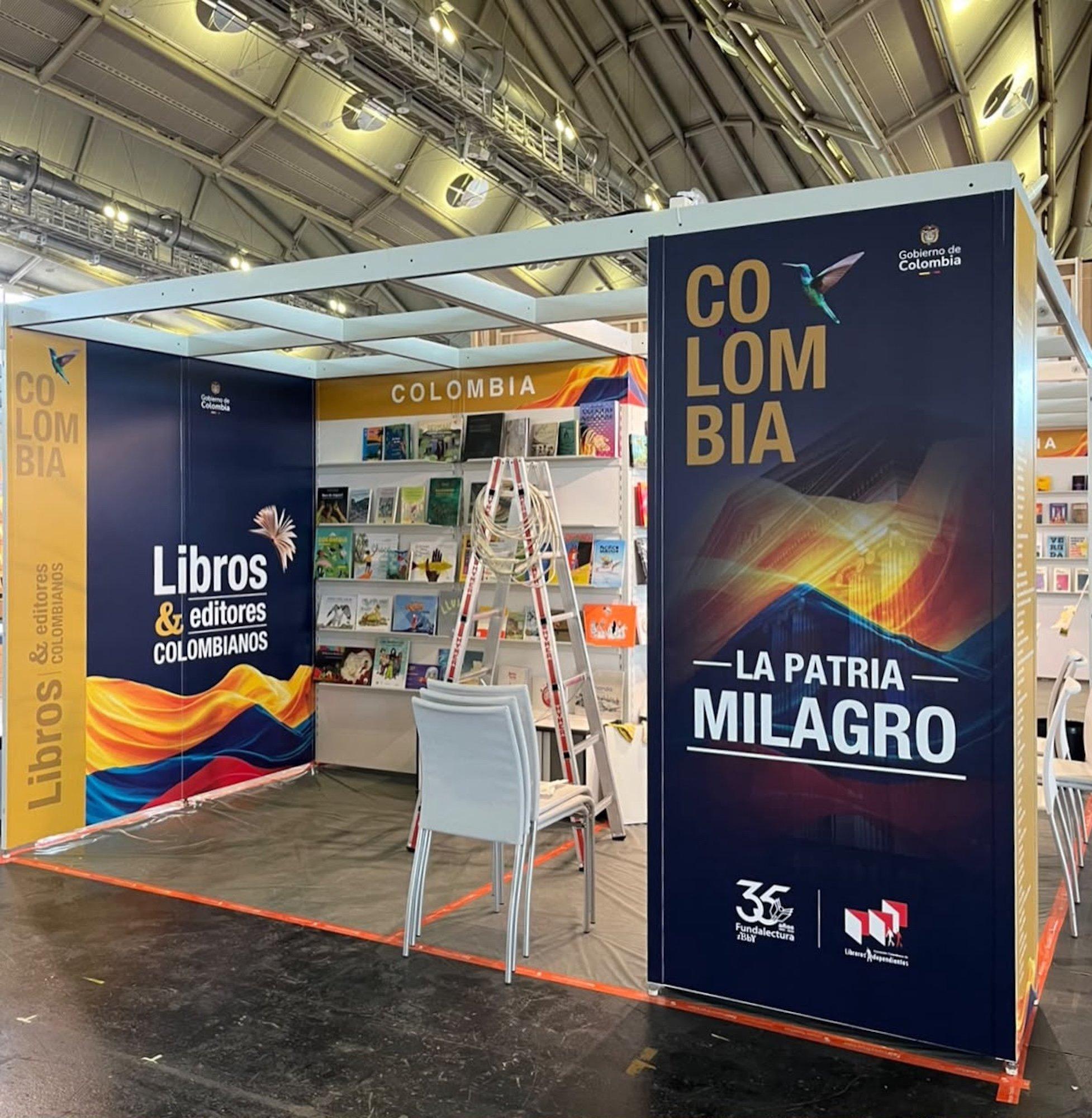 Messehalle mit Metalldach, darin ein Stand aus Metallsteckbauweise. An den Werbeflächen prangt ein simples Design mit dunkelblauem Hintergrund, unten jeweils eine stilisierte kolumbianische Fahne. Auf einer Seite steht auf Spanisch „Kolumbianische Bücher & Verleger“. Auf der anderen Seite steht „Kolumbien. Das Wundervaterland.“ Dazu ein paar Logos, manche davon von der Regierung, ein winziges stilisiertes Buch, ein winziger Kolibri. Im Stand selbst sind schon Bücher in den Auslagen, in der Mitte eine Leiter und ein Stapel Stühle.