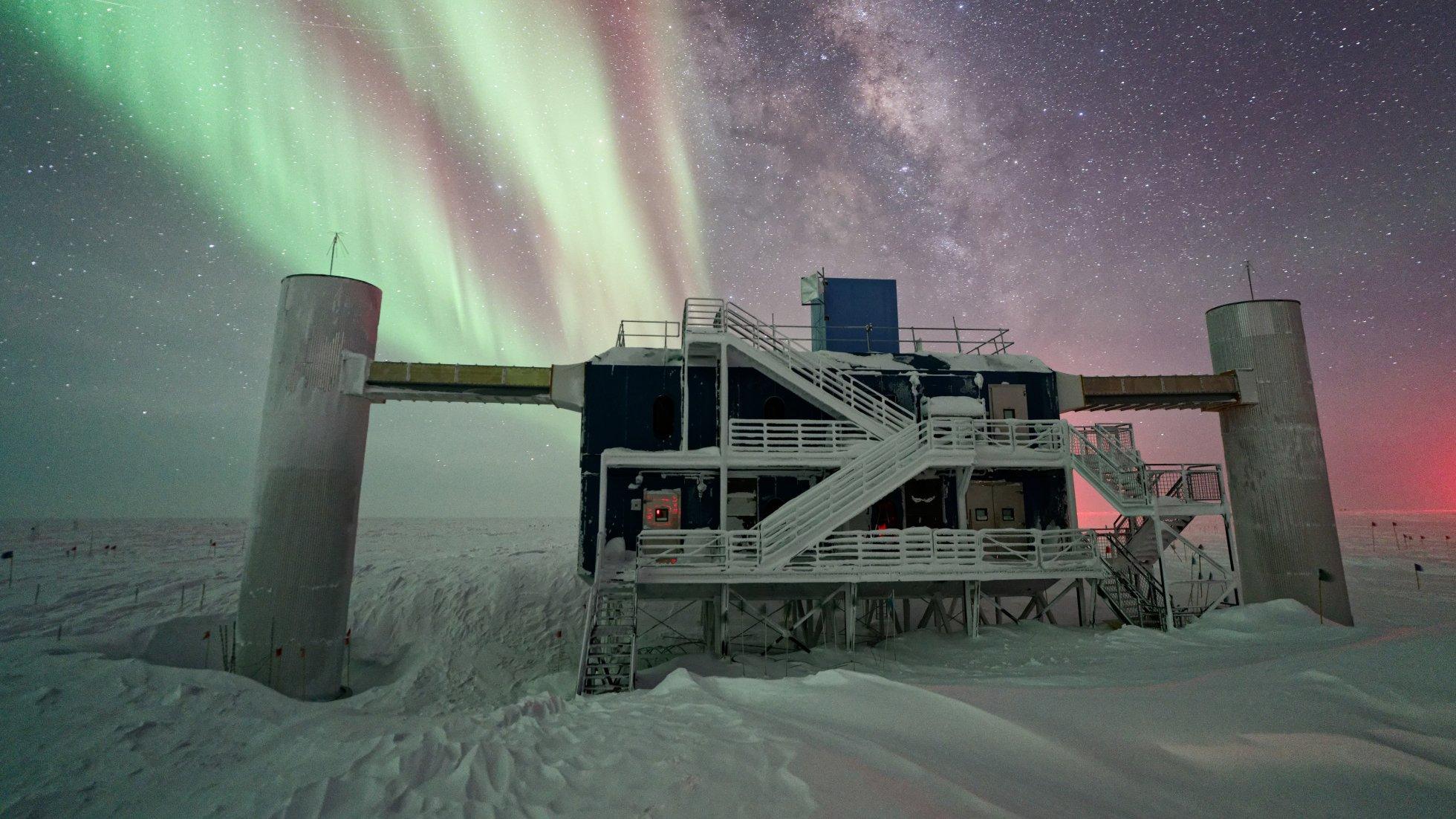 Der oberirdische Teil des Neutrinodetektors IceCube steht auf einer schneebedeckten Fläche in der Antarktis. Das Gebäude hat eine rechteckige Form mit zwei zylindrischen Seitentürmen und mehreren weißen Treppen und Geländern. Über dem Gebäude leuchten grüne und rötliche Polarlichter, die sich wellenförmig über den Himmel ziehen. Im Hintergrund ist die Milchstraße als heller, sternenreicher Streifen deutlich sichtbar.