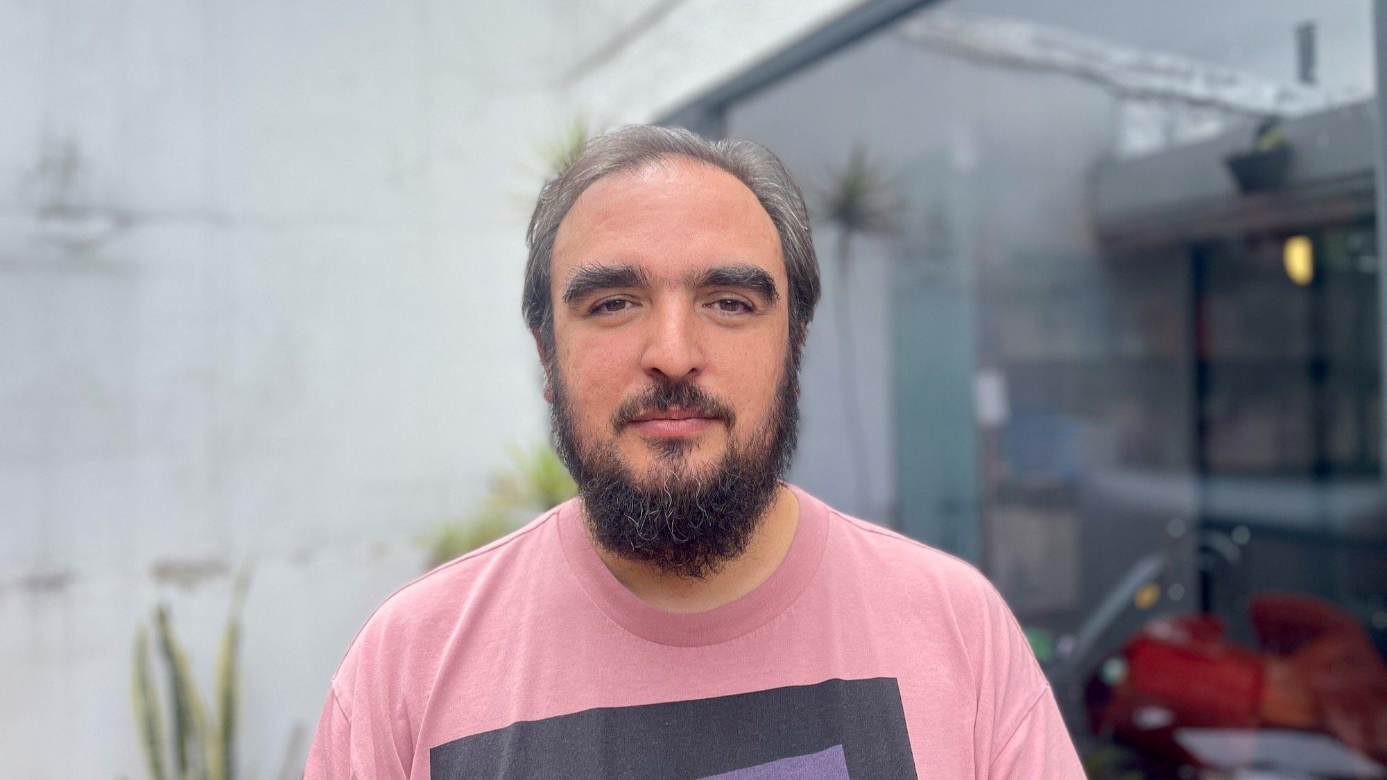 Mann um die 40, mit dunklem Vollbart, buschigen Augenbrauen und sich zurückziehendem Haupthaar blickt freundlich und ruhig in die Kamera. Er trägt ein rosa T-Shirt mit Vierecken drauf. Er steht in einem Innenhof. Hinter ihm links eine Betonwand, rechts eine Glasfront zum Buchlager.