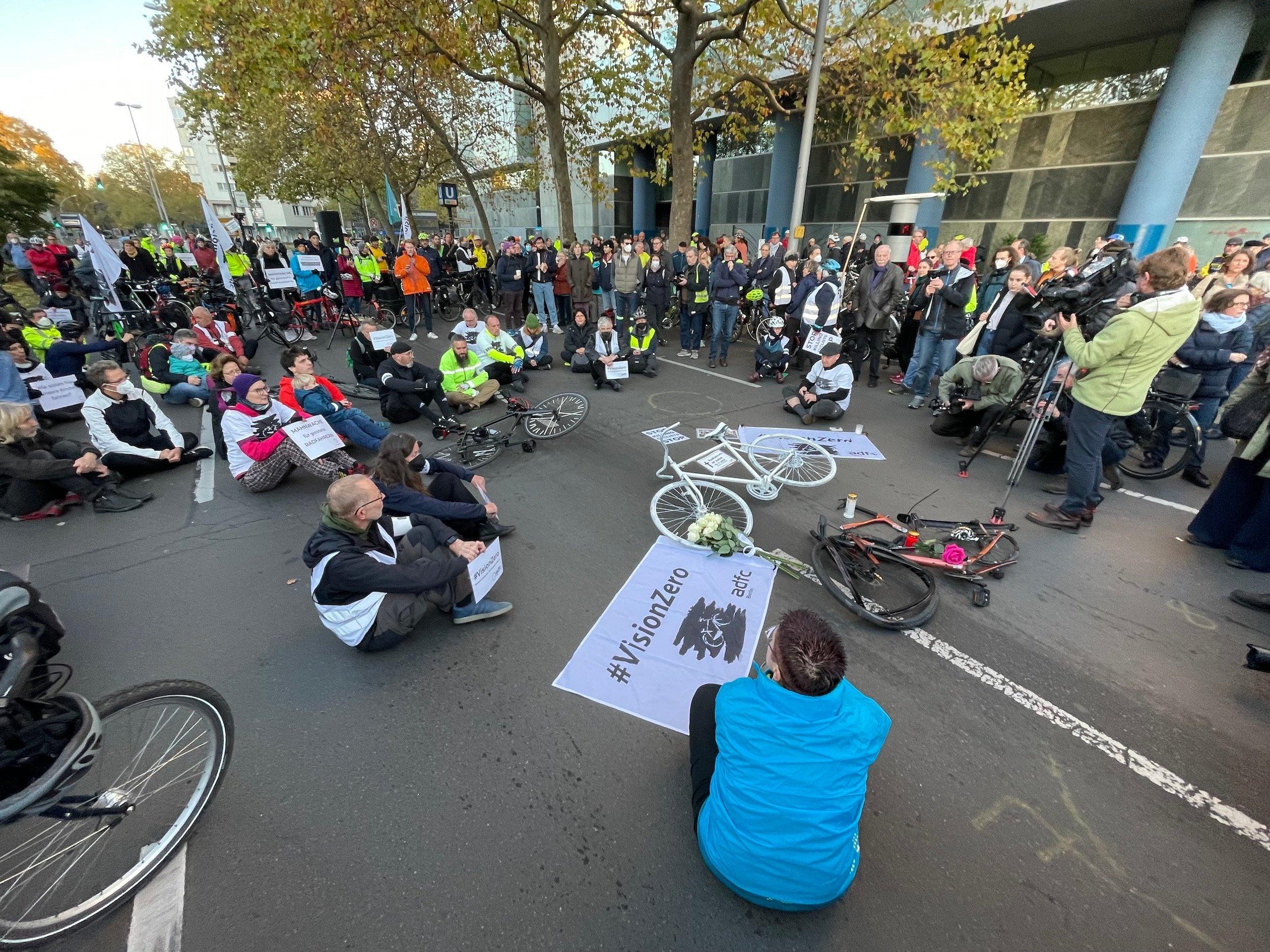 Eine Menschenmenge versammelt sich um ein komplett weiß bemaltes Fahrrad, das mitten auf der Straße liegt. Daneben ein Banner #VisionZero für das Ziel, dass es Null Verkehrstote gibt.