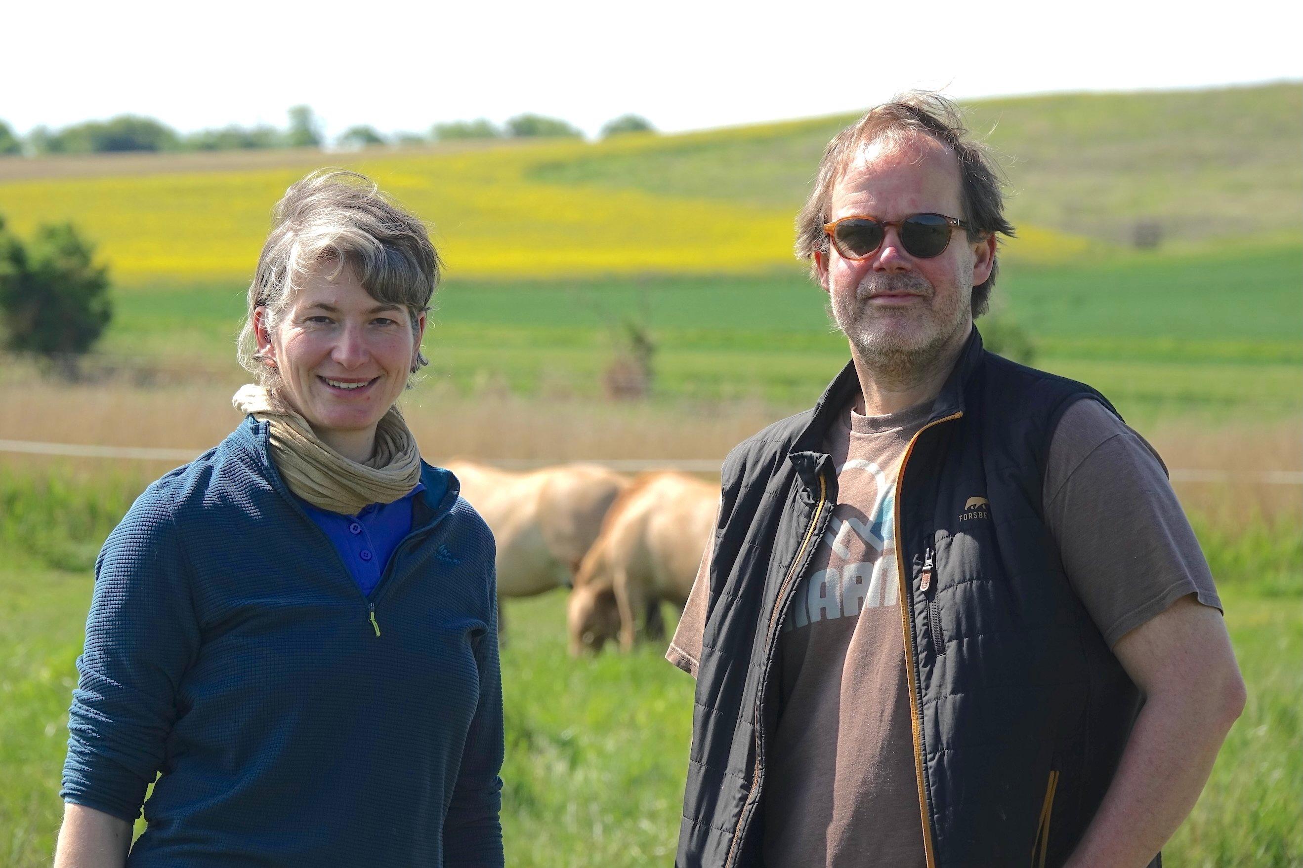 Frau und Mann in grüner Landschaft, im Hintergrund Pferde.
