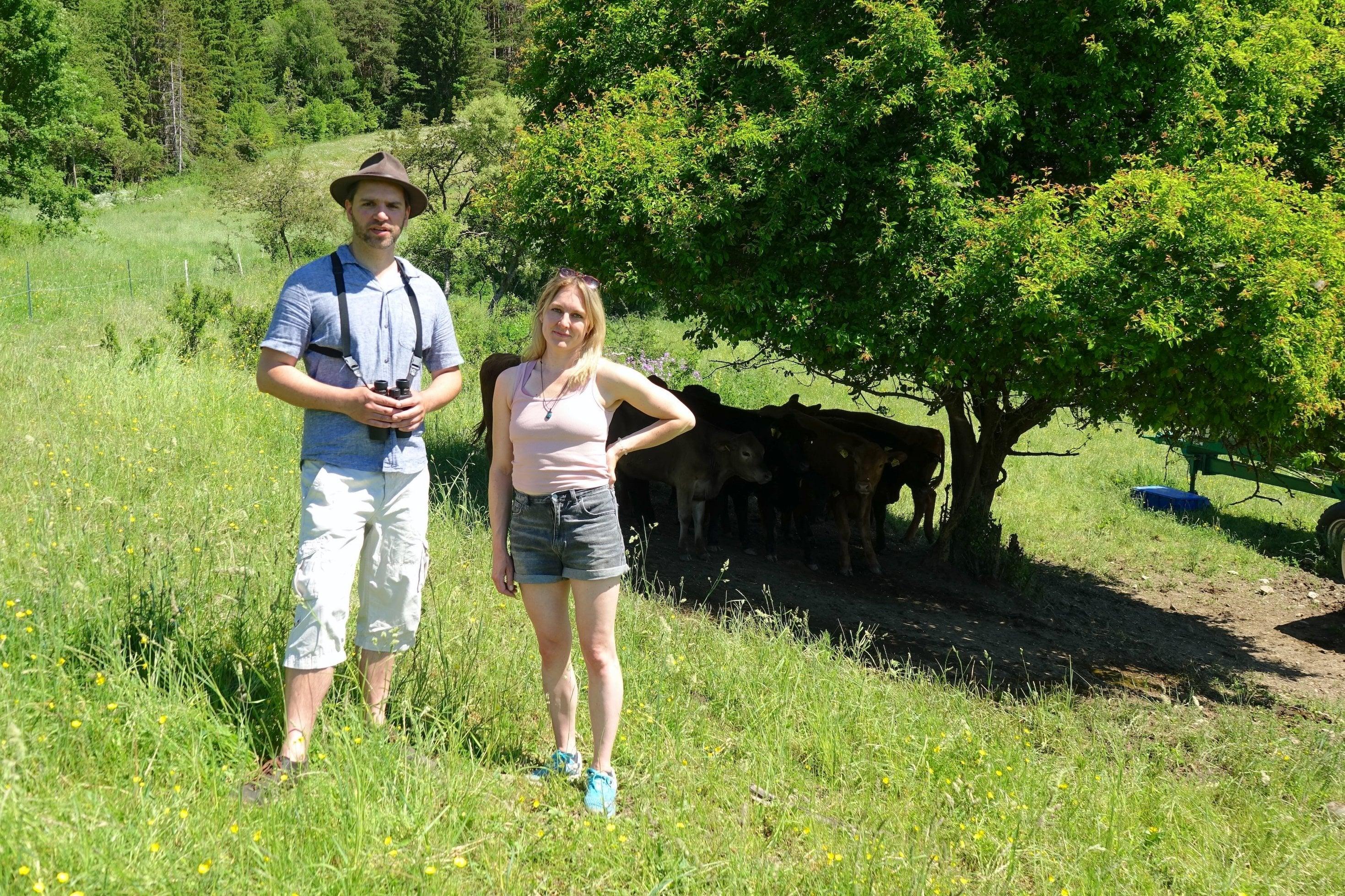 Mann mit Hut und Frau in sportlicher Kleidung vor jungen Rindern in grüner Landschaft.