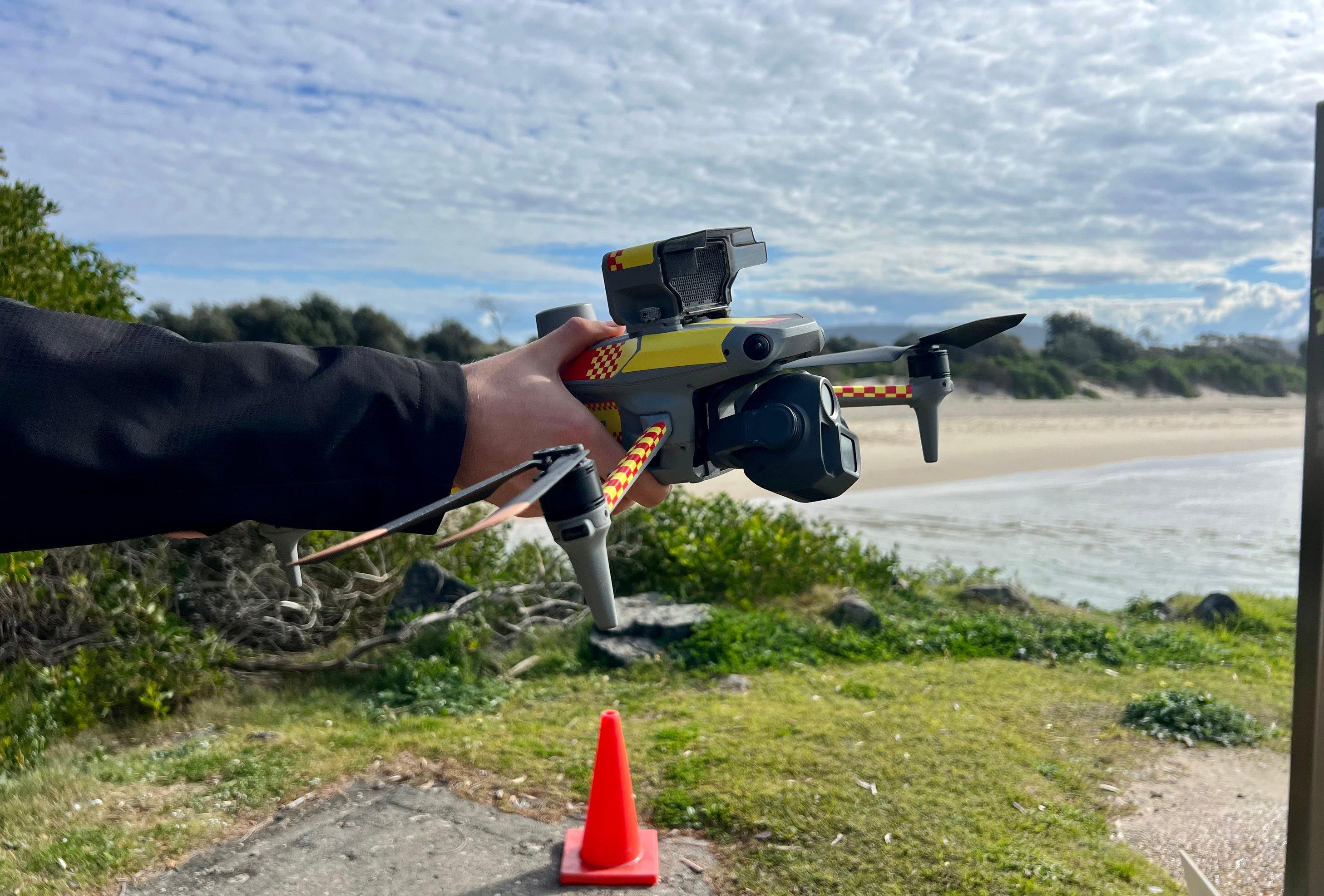 Ein Drohnenpilot hält eine Drohne, die einen Lautsprecher hat, die Lautsprecher an können Schwimmer und Surfer draußen im Meer vor Gefahren warnen.