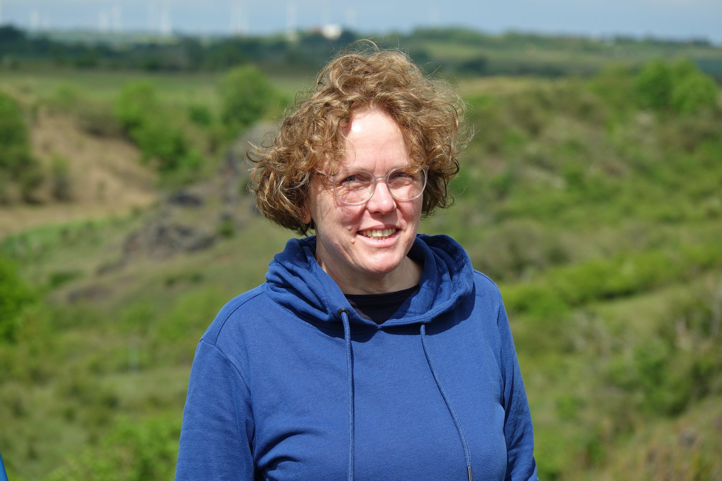 Freundlich schauende Frau mit roten Haaren vor weiter Landschaft.