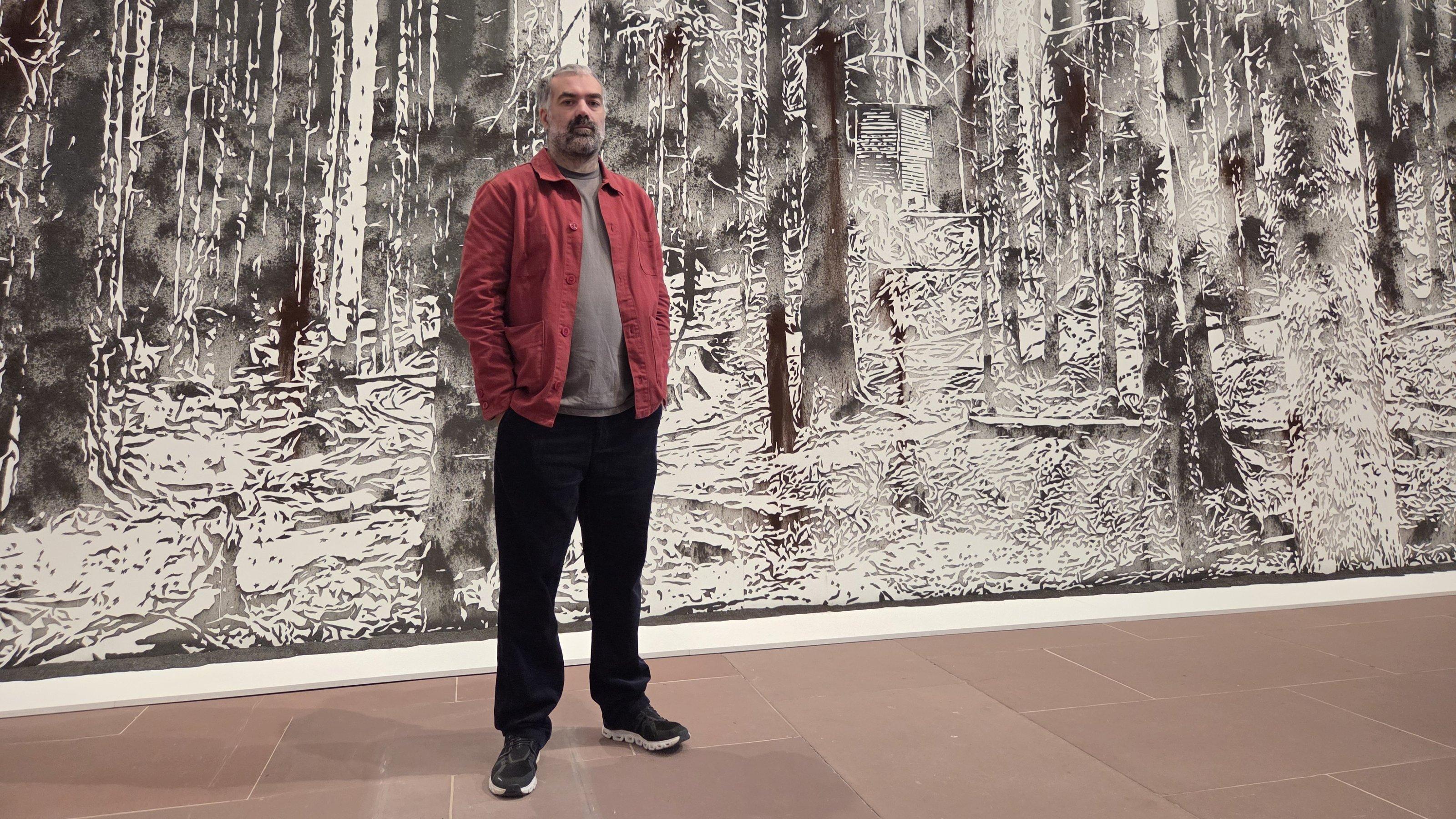 Ein bärtiger Mann mit roter Leinenjacke steht vor einer wandfüllenden Leinwand.