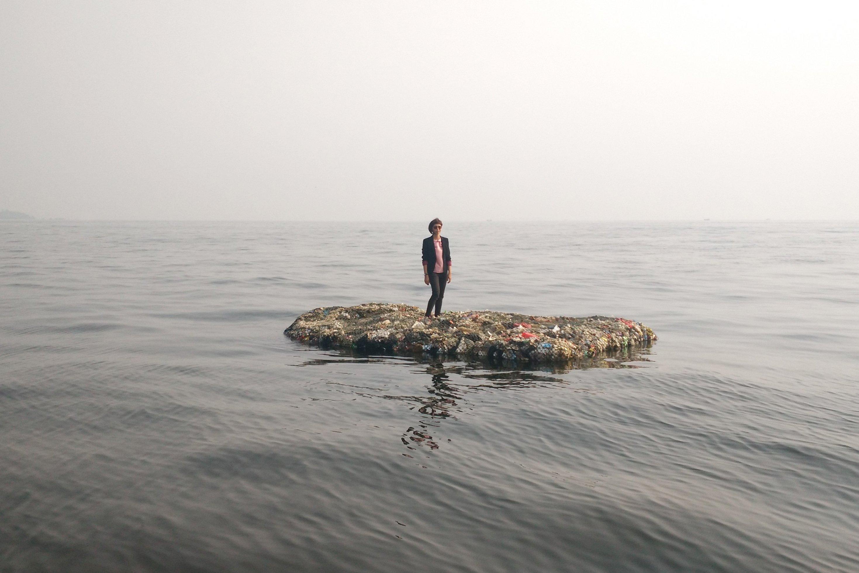 Ein Floß aus Plastikmüll treibt auf ruhiger See. Darauf steht die Künstlerin Tita Salina.