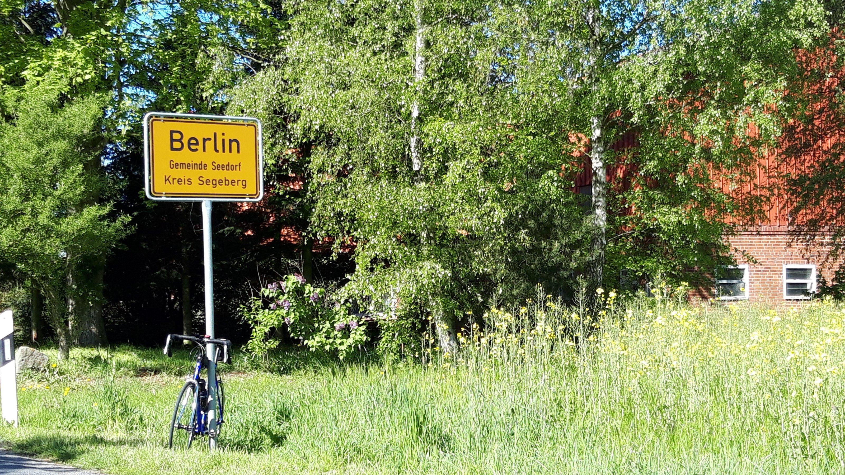 Ortsschild von „Holstein-Berlin“, an dem das Rad des RadelndenReporters lehnt.