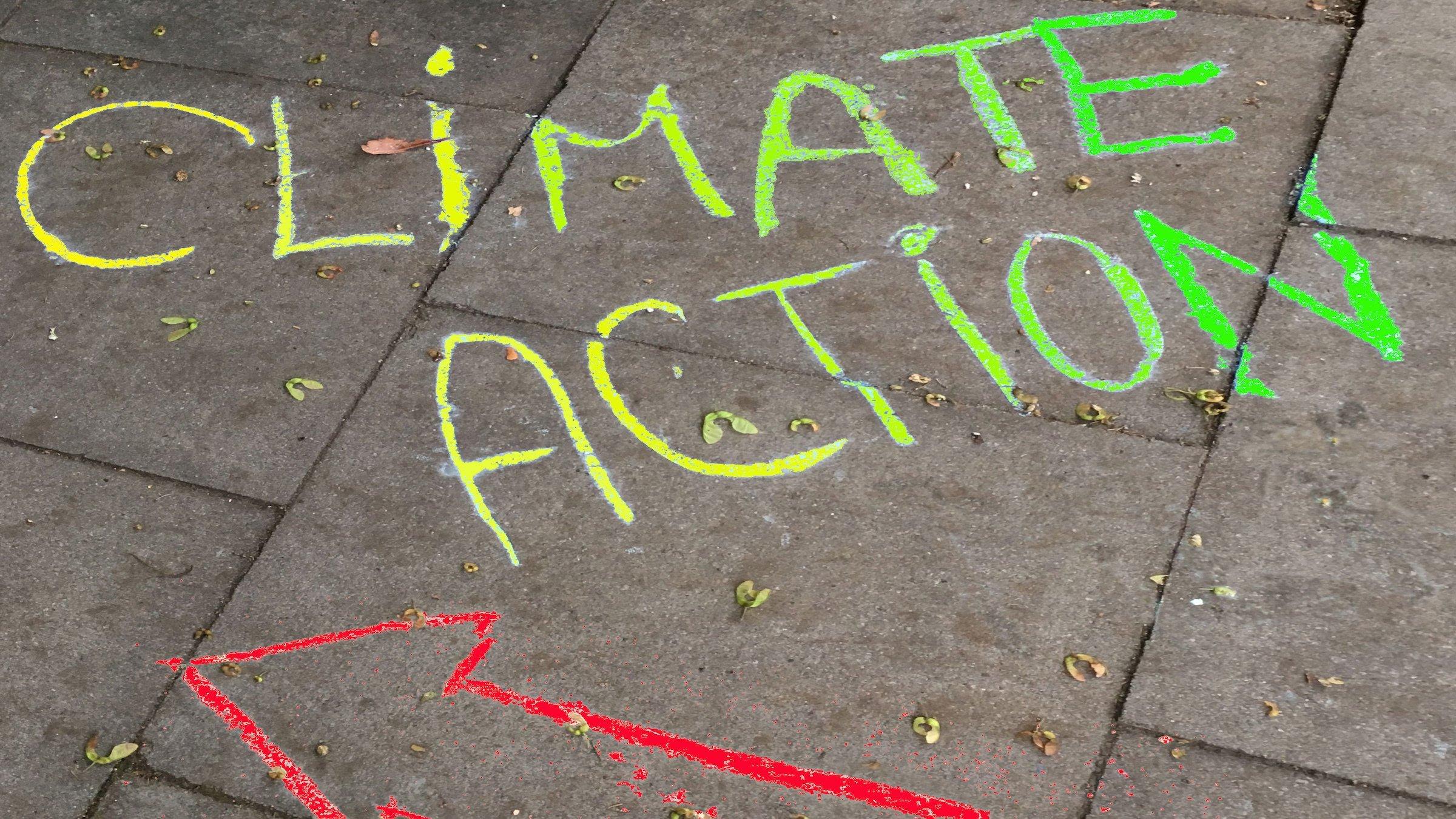Die Worte Climate Action sind mit Kreide auf das Pflaster eines Gehweges beschrieben. Die Buchstaben sind von C bis E und von A bis N mit Farben von gelb bis grün eingefärbt. Darunter zeigt ein roter Kreide-Pfeil nach links.