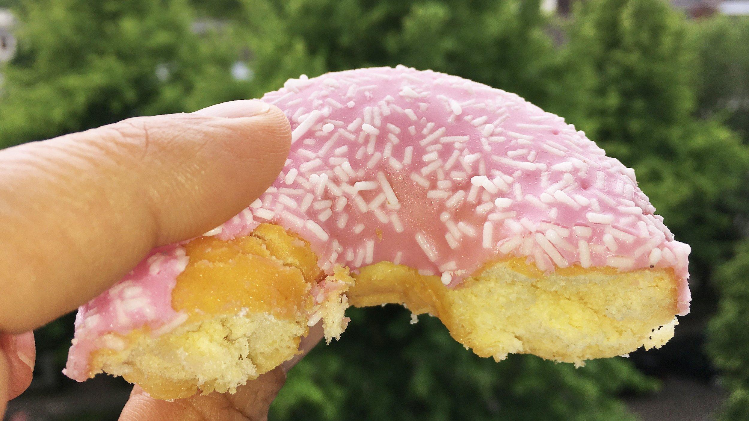Zwei Finger halten einen angebissenen Donut vor die Kamera, der mit rosa Zuckerguss verziert ist. Im Hintergrund sind unscharf Bäume zu erkennen.