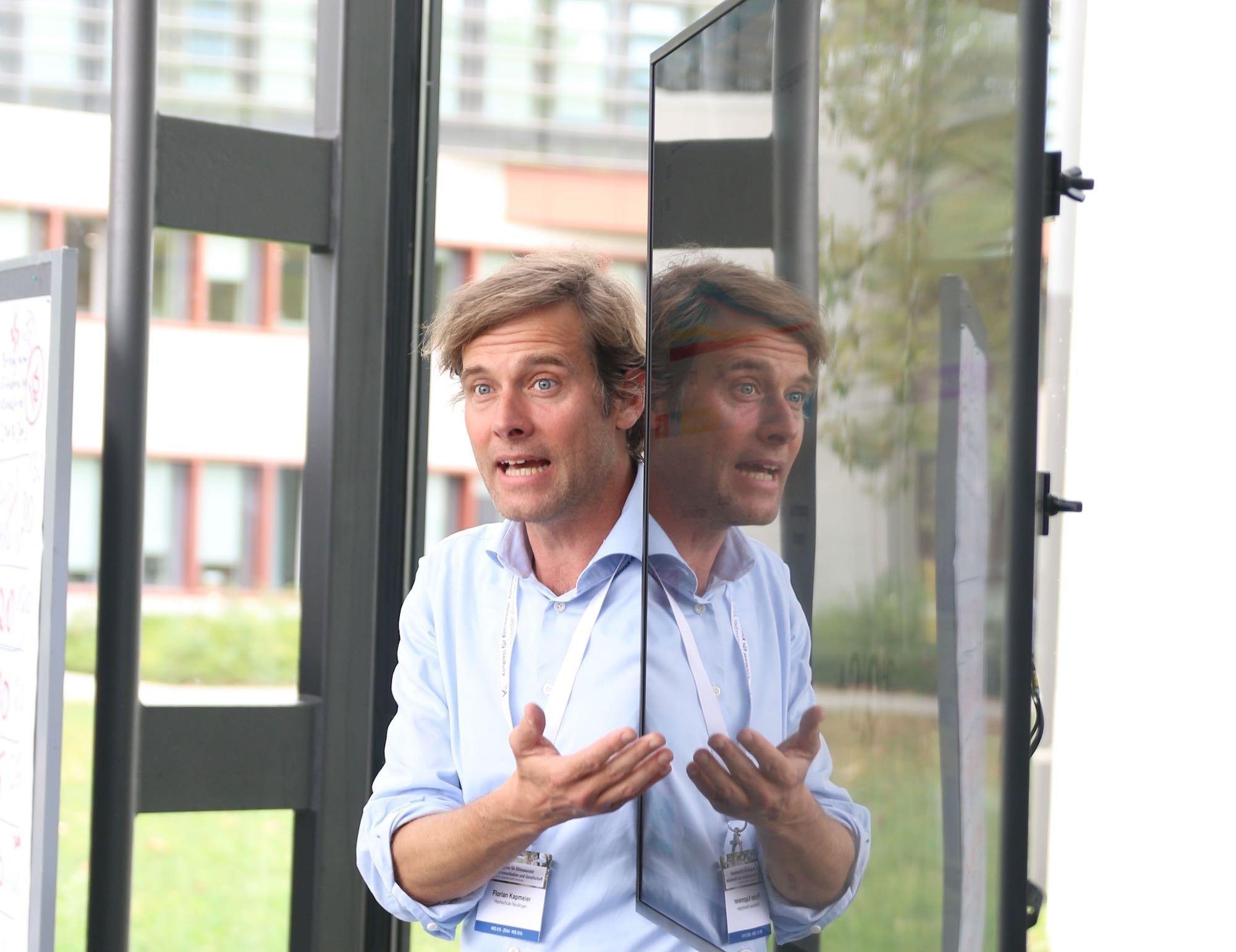 Florian Kapmeier präsentiert das En-ROADS-Modell bei einer Konferenz in Karlsruhe. Er steht vor einem Bildschirm und gestikuliert, sein Oberkörper spiegelt sich in der Glascheibe.