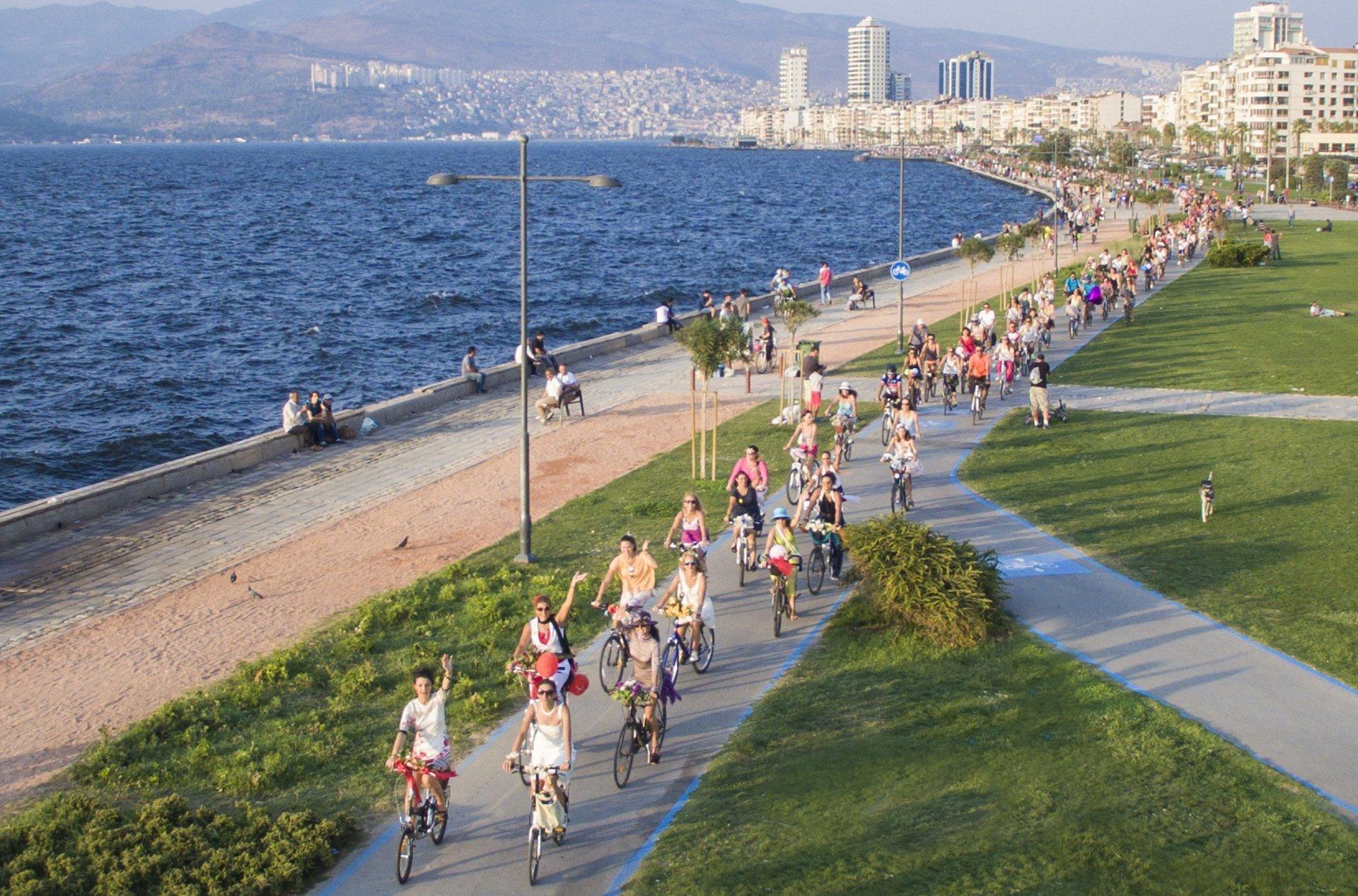 In der Hafenstadt Izmir führt ein Radweg am Meer entlang. Aus der Vogelperspektive sind die Teilnehmerinnen der Ausfahrt als farbenfrohe Punkte auf dem hellen Radweg zu sehen. Linkerhand liegt das Meer.