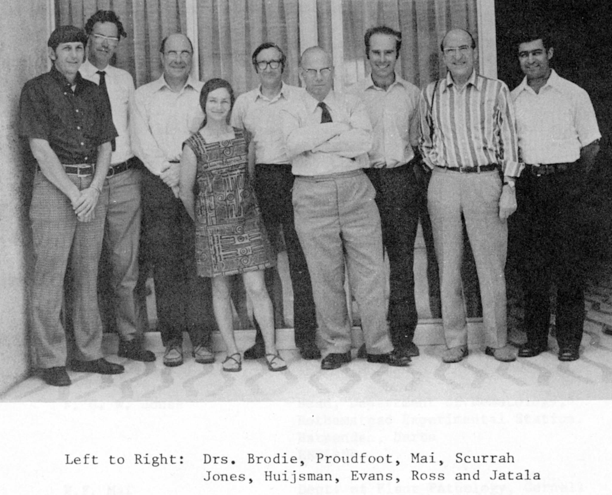Schwarzweissfoto aus dem Jahre 1976. Gruppenbild von acht weissen Männern verschiedensten Alters. In der ersten Reihe eine junge, kleine, zierliche Frau.