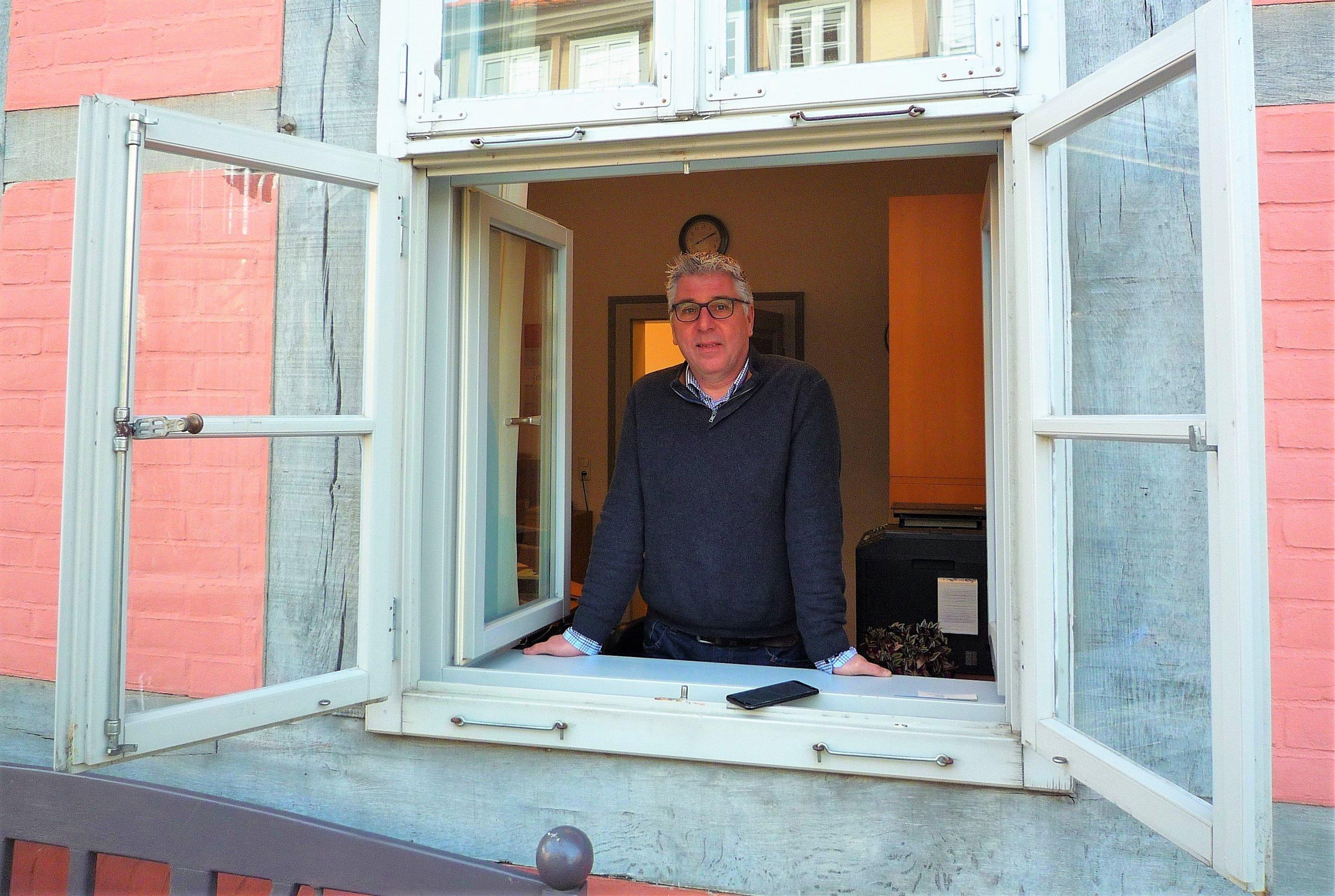 Mann mit Brille und Pullover stützt sich von innen auf das Brett des Fensters, dessen Flügel weit nach außen offenstehen.