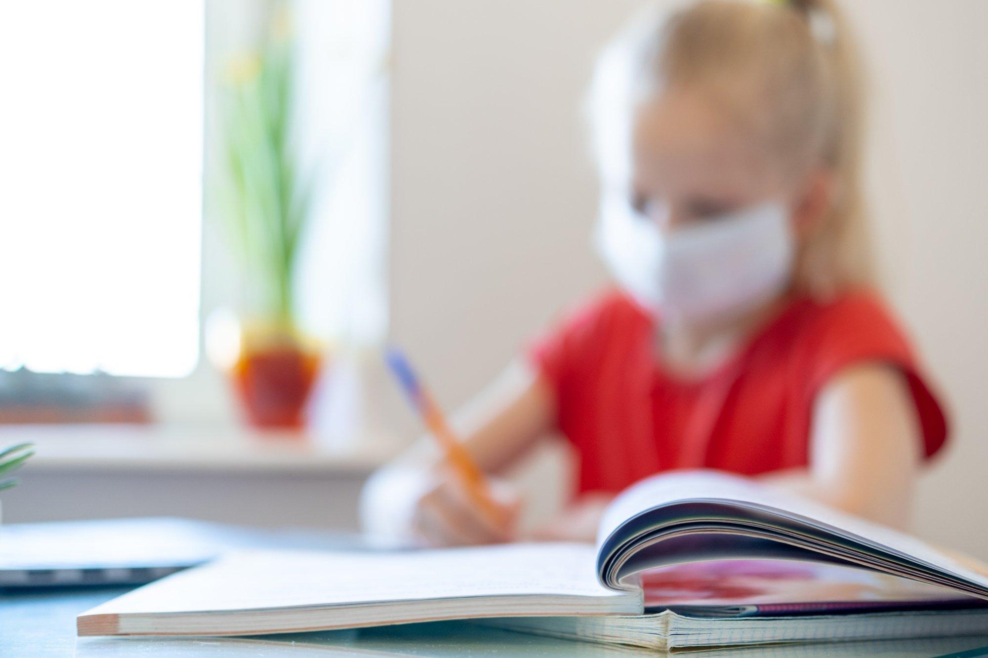 Ein kleines Mädchen sitzt mit Maske an einem Schreibtisch und macht Aufgaben in einem Schulheft.