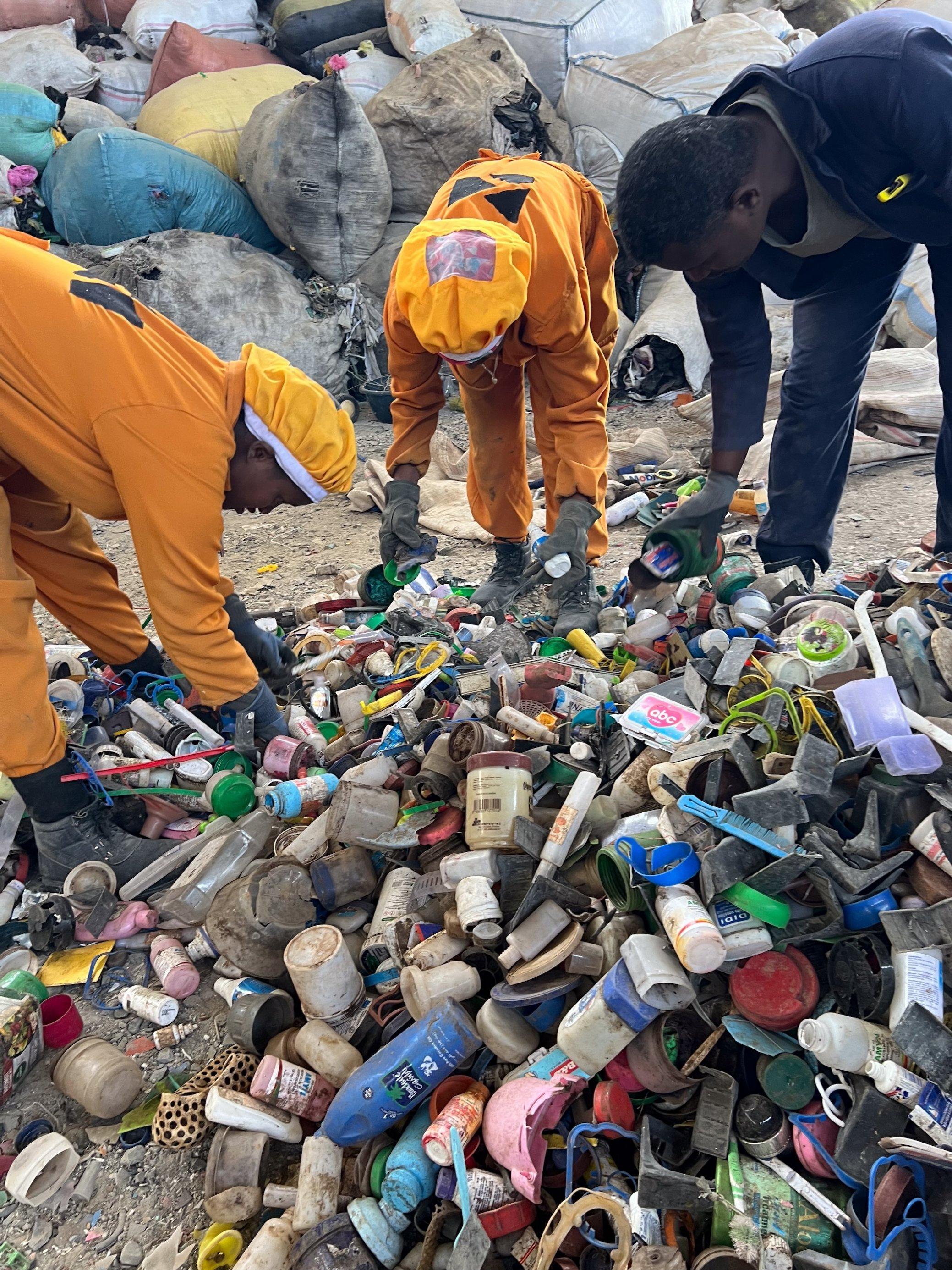 Zwischen Bergen aus Plastikmüll bücken sich drei Menschen, um die Teile zu sortieren. Zwei Frauen tragen orangene Overalls und einen Haarschutz, ein Mann einen dunkelblauen Overall.