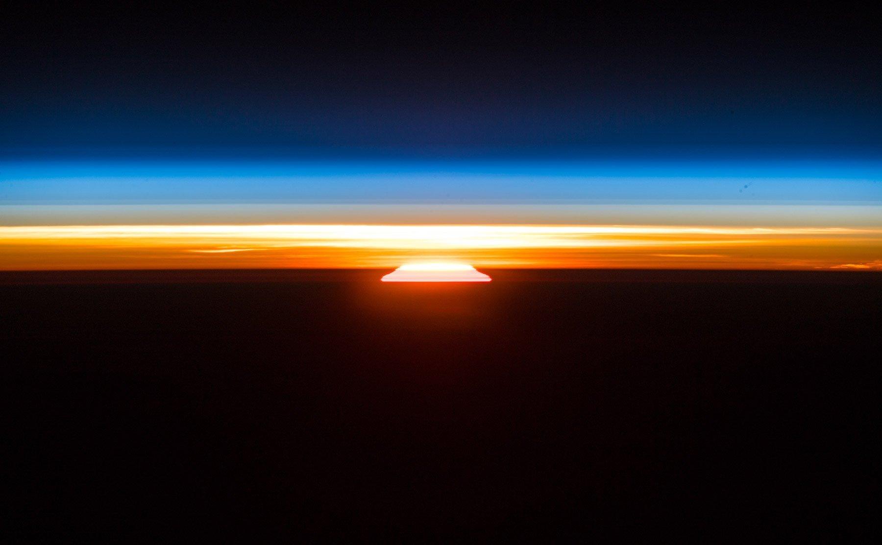 Ein Sonnenaufgang, vom Erdorbit aus vom deutschen Raumfahrer Alexander Gerst fotografiert. Über der aufgehenden Sonne die dünne, orange-blaue Erdatmosphäre, darüber die Schwärze des Weltalls.