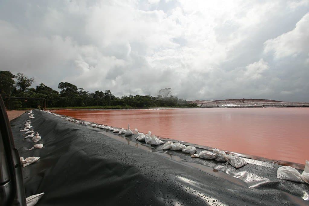 Eine Totale zeigt ein Wasserbecken mit rostrotem Wasser auf dem Alunorte-Gelände. Im Vordergrund sieht man eine schwarze Begrenzung, vermutlich aus Plastikplanen.
