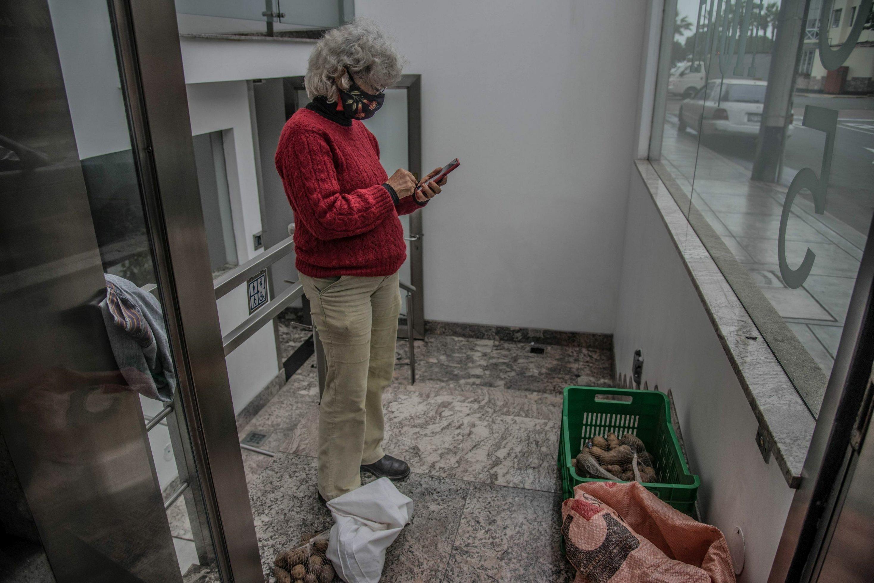 Eingangsbereich eines modernen Hochhauses. Eine Frau mit beiger Cordhose und rotem Pulli, weissen Locken und Mundschutz, tippt etwas in einen Taschenrechner. Vor ihr auf dem Boden eine Kiste mit Kartoffeln.
