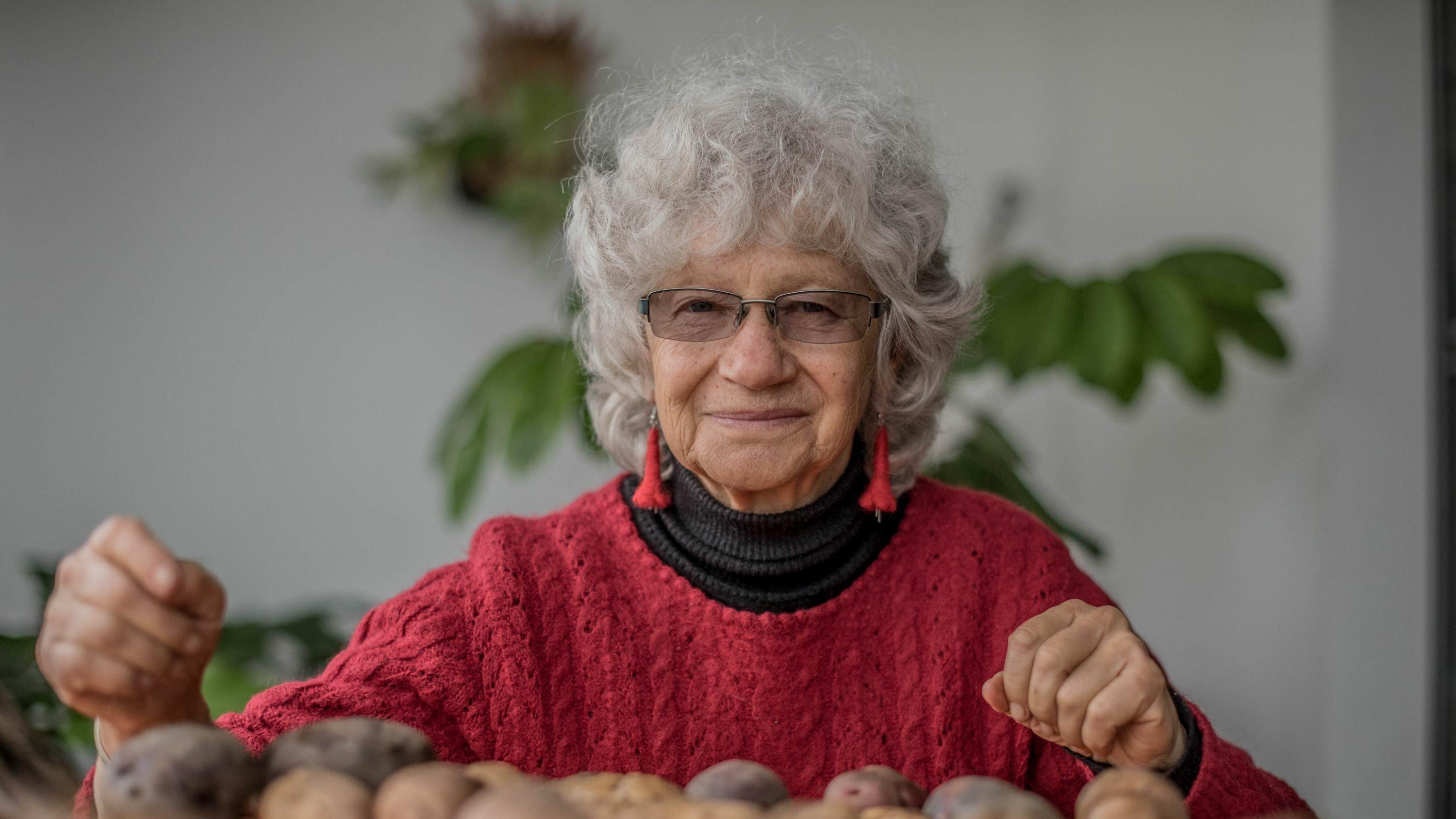 Weißhaarige Frau mit runzligem Gesicht und rotem langärmligen Pullover blickt in die Kamera, vor sich auf dem Tisch liegen mehrere Kartoffeln.