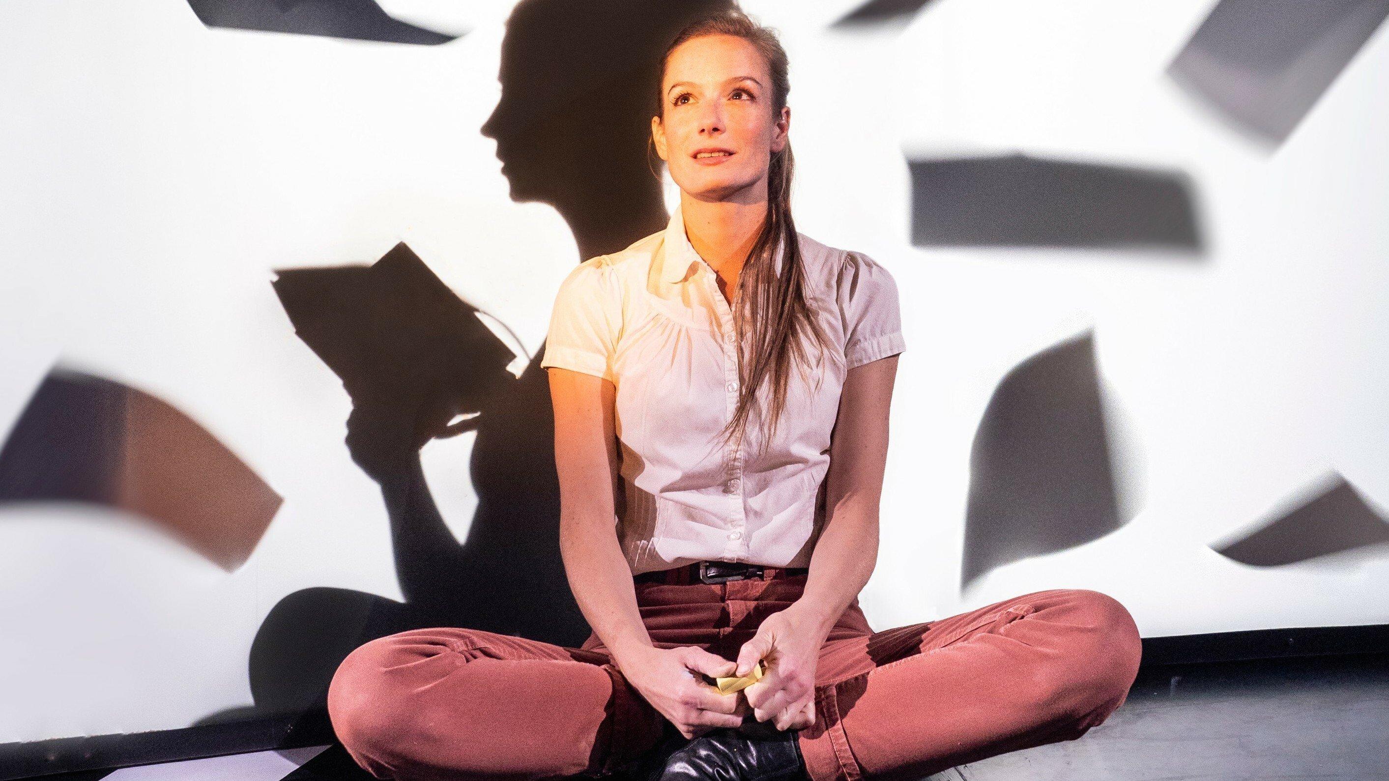 Eine junge Frau mit langen Haaren sitzt nachdenklich im Schneidersitz vor einer weißen Wand. Dahinter sitzt wie ein Schatten eine weitere Schauspielerin. Um sie herum fliegen Notizblätter. Das Bühnenbild von Ansgar Prüwer spiegelt, was Anne Frank sich vorstellt.