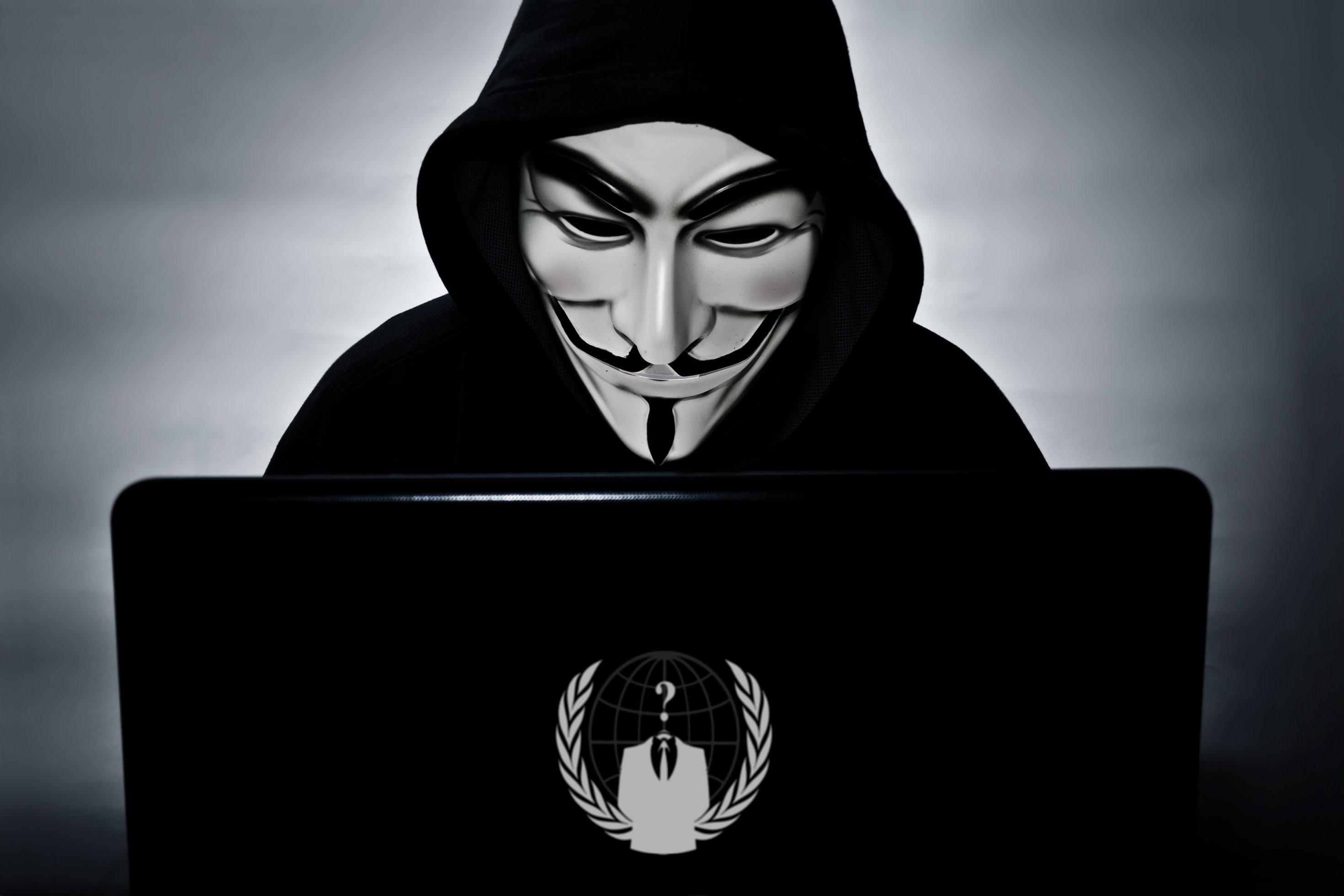 Ein Mensch mit einer weißen Maske mit Bart und schwarzem Kapuzenpulli sitzt vor einem Laptop, auf dem das Logo des Hacker-Kollektivs „Anonymous“ abgebildet ist.