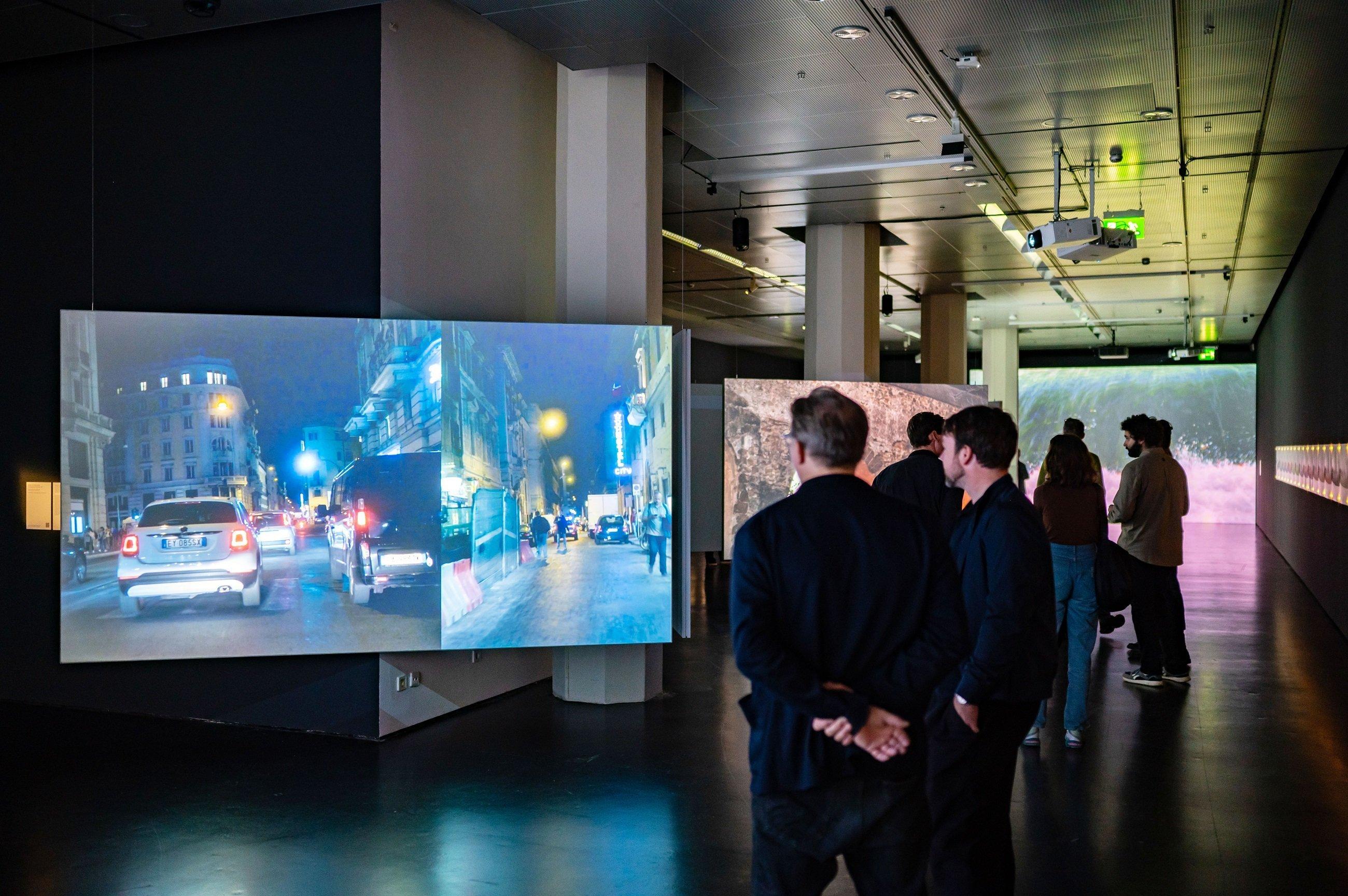 Blick in die Ausstellung: ein dunkler, langgestreckter Raum. Große Flatscrees sind zickzackförmig angeorndet. Davor Besucher der Schau.