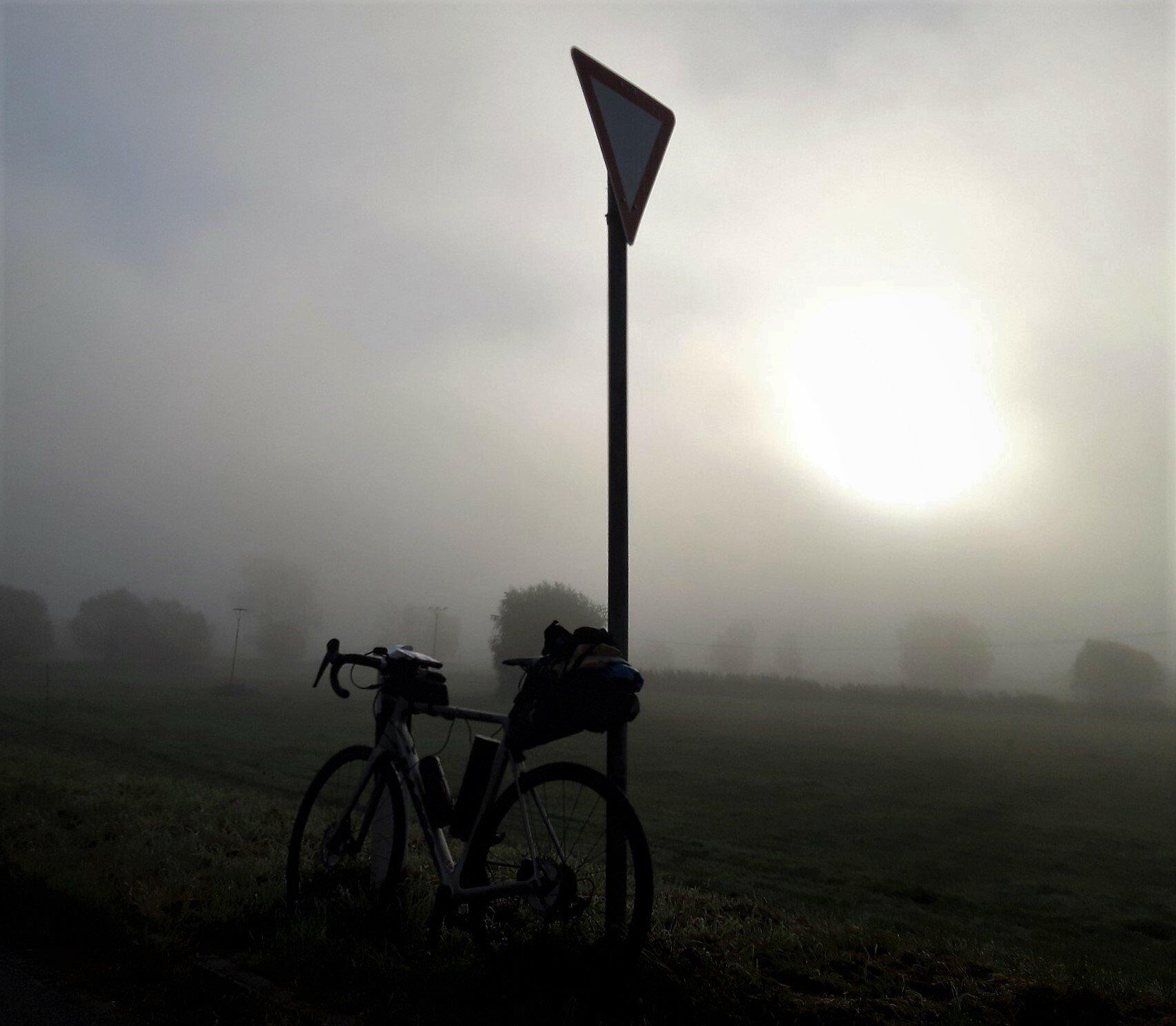 Das Fahrrad lehnt an einem „Vorfahrt-achten“-Schild. Straße, Felder und Baumgruppen sind von Nebel umhüllt, durch den sich eine grellweiße Sonne zu brennen versucht.