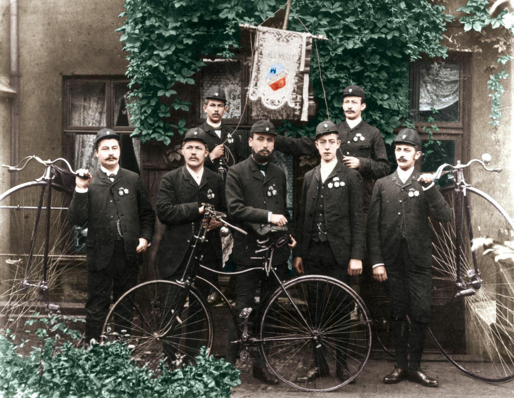 Sieben Männer in Sonntagstracht posieren mit einem Fahrrad, auf dessen Sattel Harro Feddersen, im Zentrum stehend, eine Hand auflegt. In der hinteren Reihe stützen zwei Männer eine Vereins-Standarte.