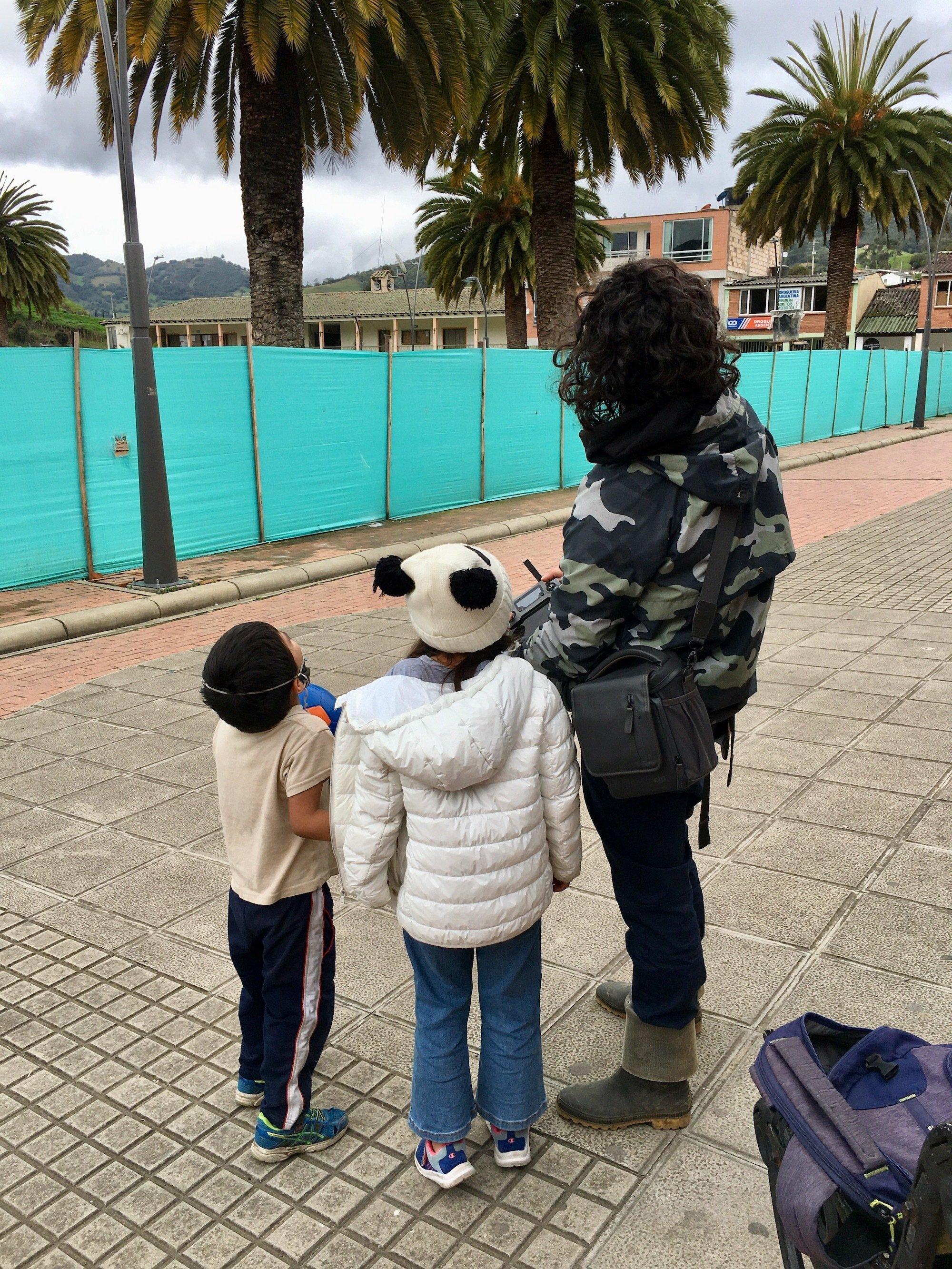Ein kleiner Junge, ein Mädchen mit Pandamütze und ein Mann mit lockigem Haar, Umhängetasche und Fernbedienung in der Hand blicken nach oben. Alle sind nur von hinten zu sehen.