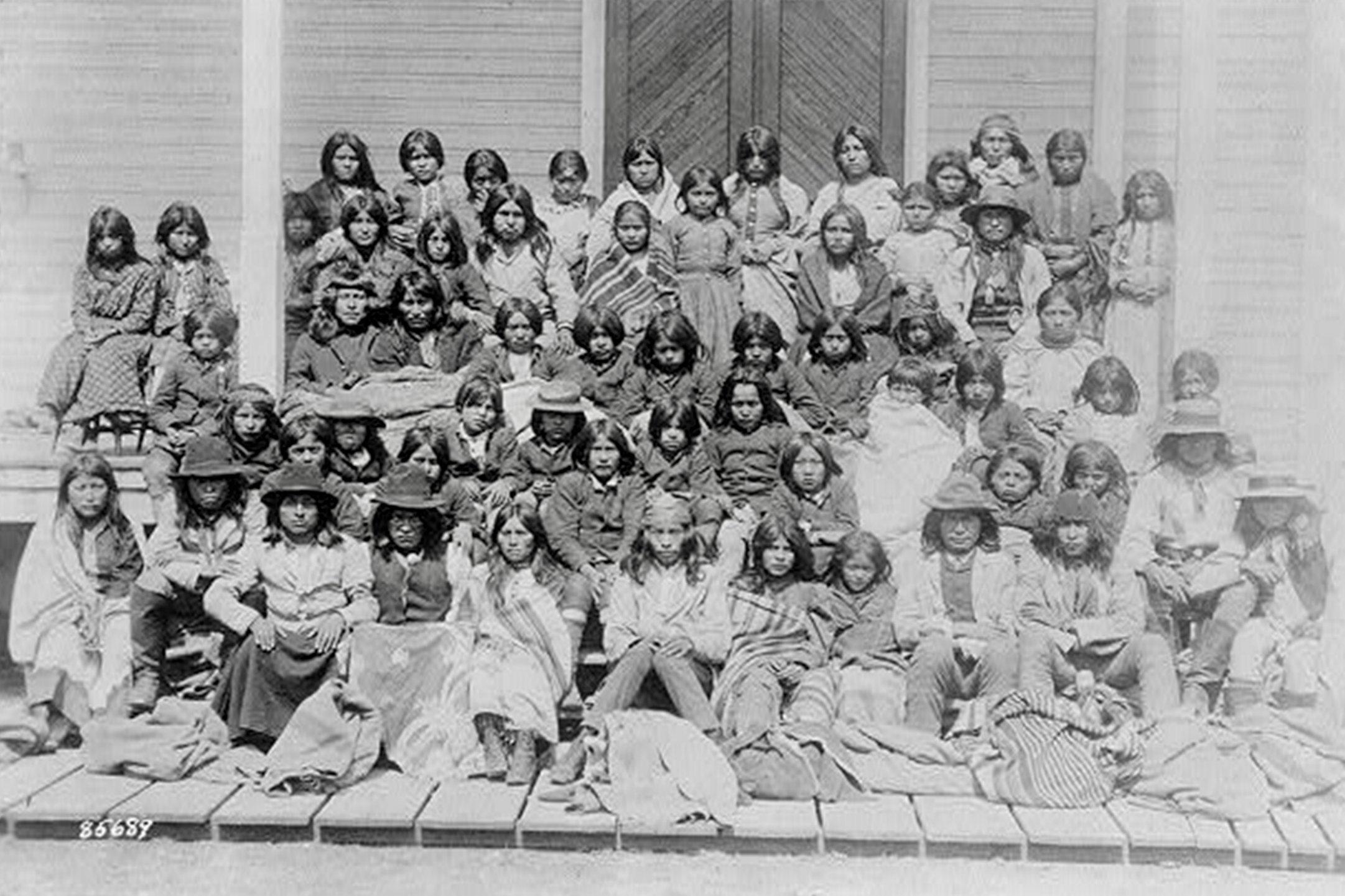 Eine Gruppe von etwa 50 Kindern posiert bei der Ankunft im Internat in Carlisle Pennsylvania in fünf Reihen sitzend auf den Treppen des Gebäudes. Die meisten haben langes dunkles Haar, manche tragen Decken über ihrer Kleidung. Sie schauen direkt in die Kamera