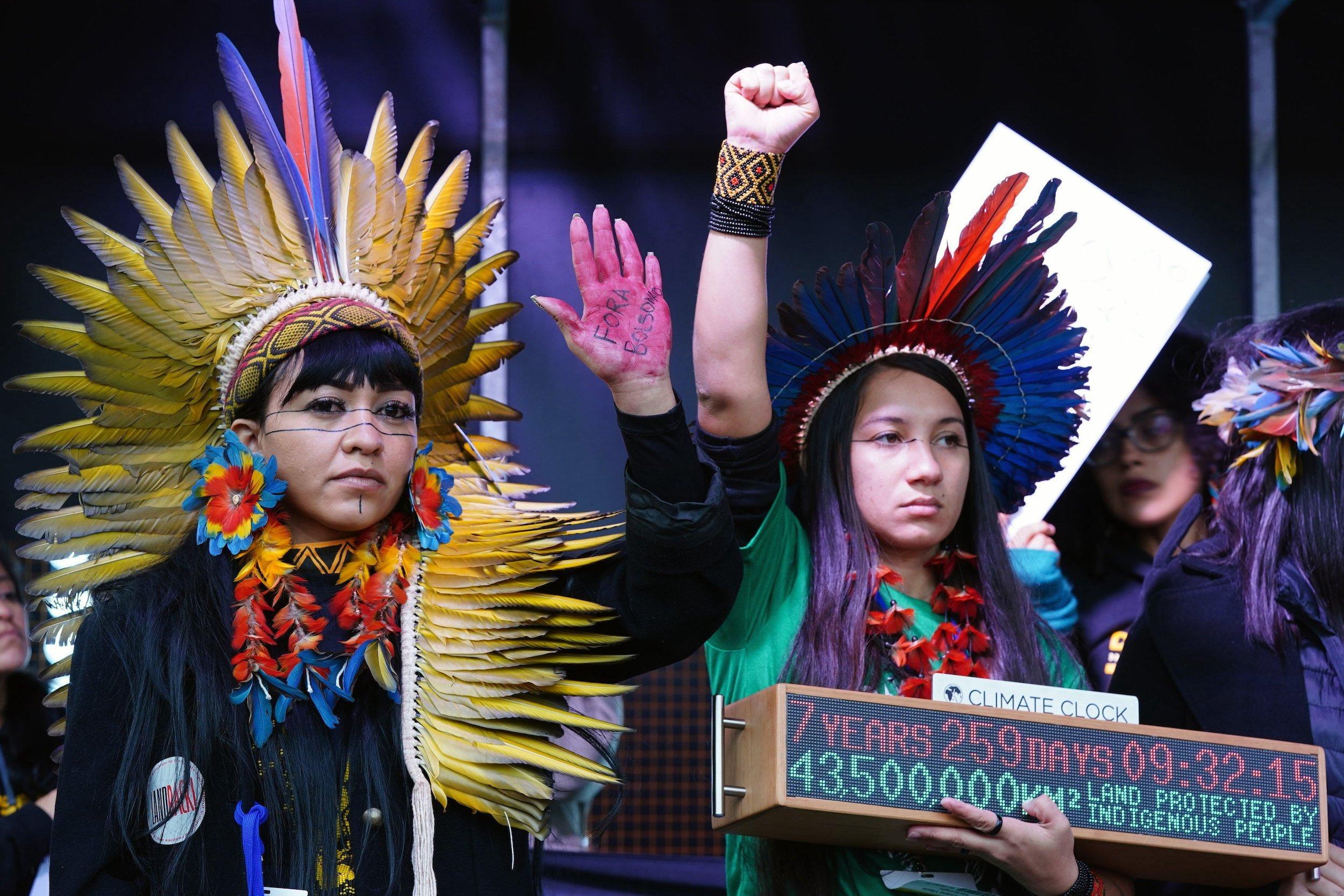 Zwei junge Frauen in indigener Tracht mit geballten Fäusten auf einer Bühne.