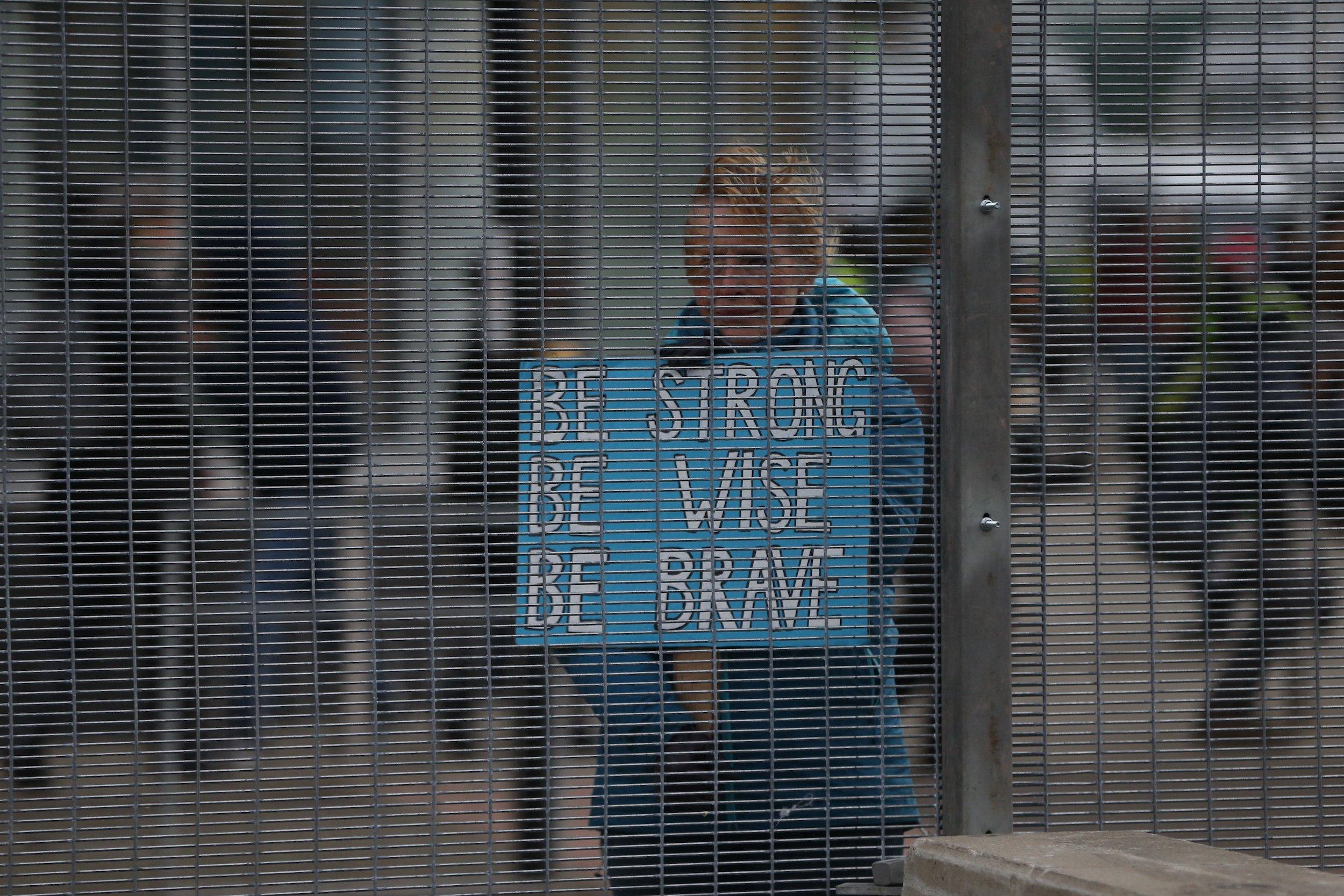 Eine Demonstrantin hinter einem Gitter mit dem Schild „Be strong, be wise, be brave“.