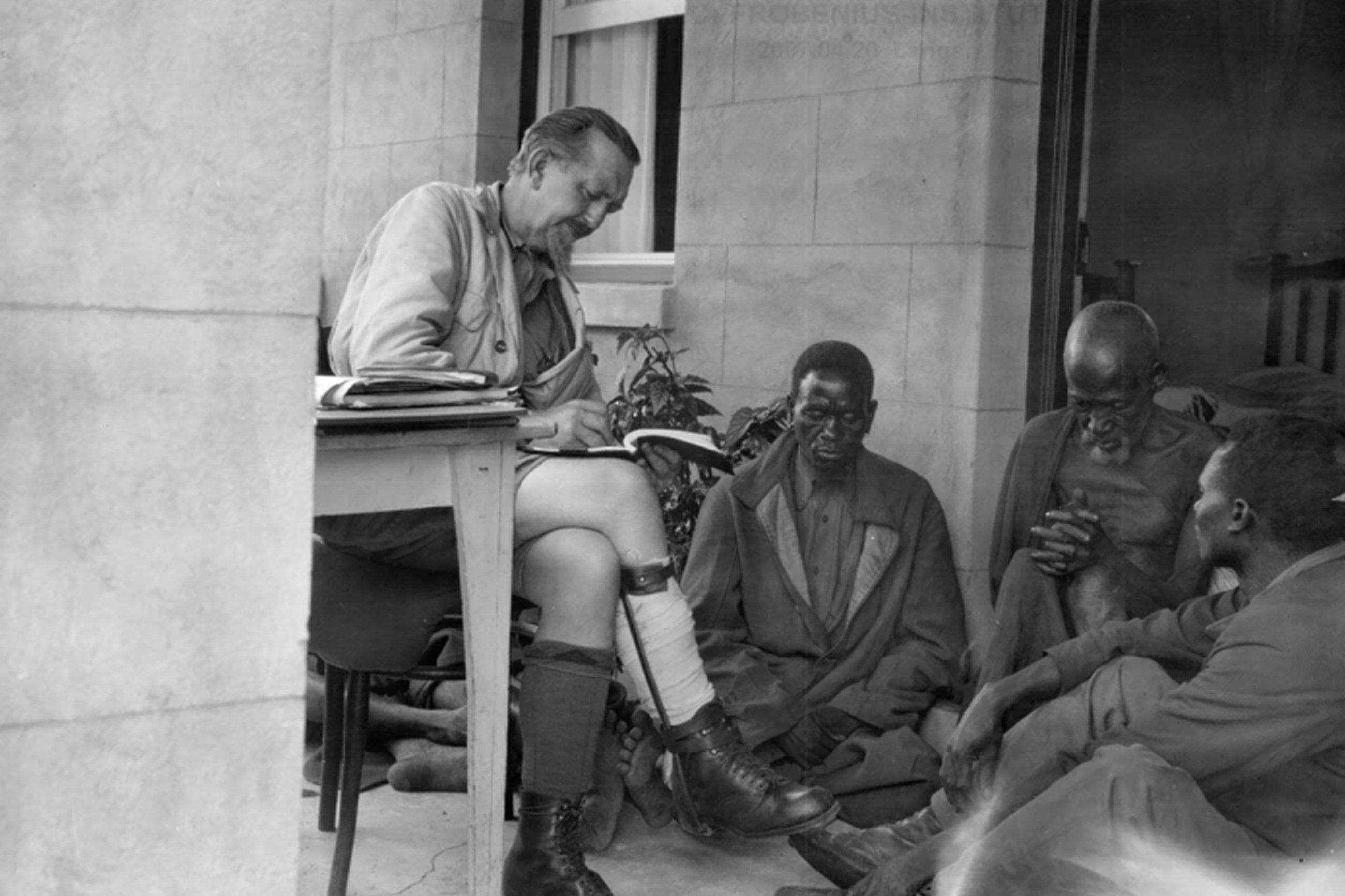 Ein weißer, älterer Mann sitzt mit Stift und Papier in der Hand vor drei auf dem Boden sitzenden Männern.