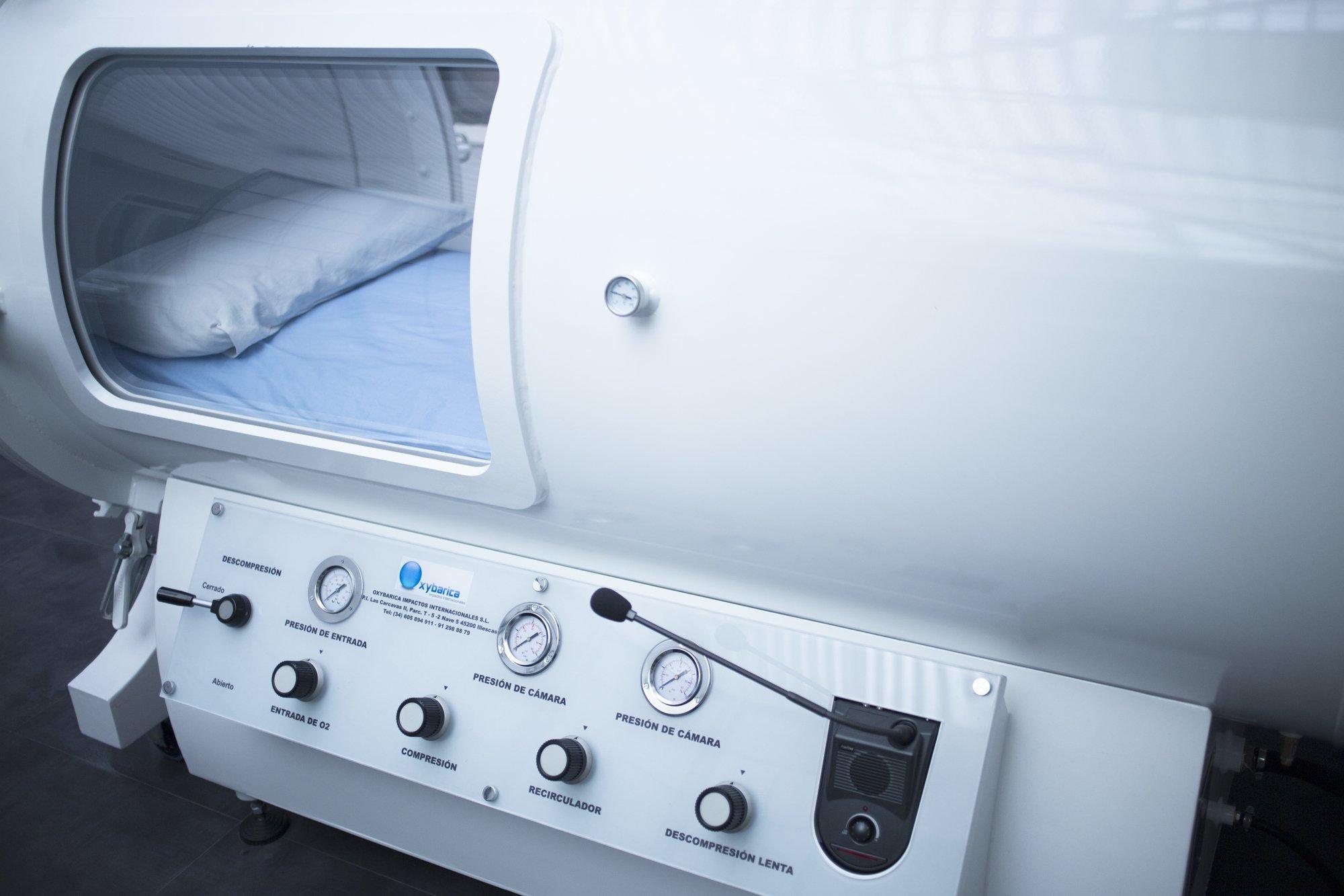 Eine Sauerstoff-Druckkammer in Form einer großen, weißen Kapsel mit Instrumenten zur Bedienung. Ein Fenster offenbart den Blick ins Innere der Kammer: Patient:innen können sich zur Behandlung hier auf eine Matratze und ein Kopfkissen legen.