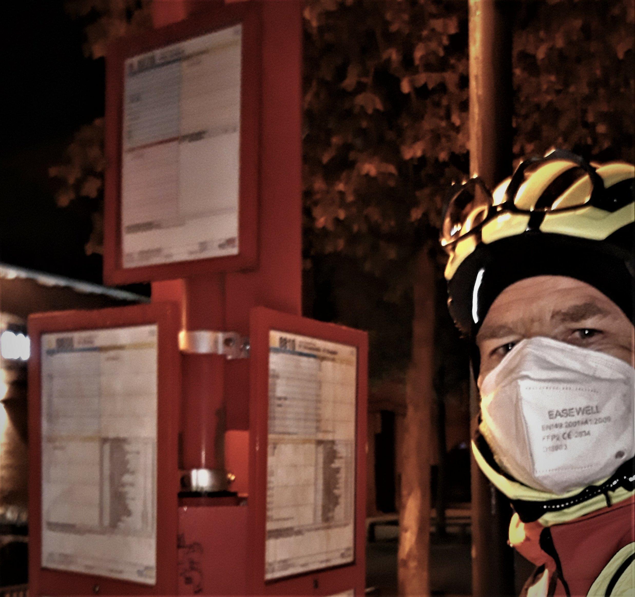 Selfie des mit Helm, Anti-Corona-Maske und Kälteschutzutensilien vermummten Reporters vor einem roten Pfosten mit rot umrandeten Busfahrplänen.