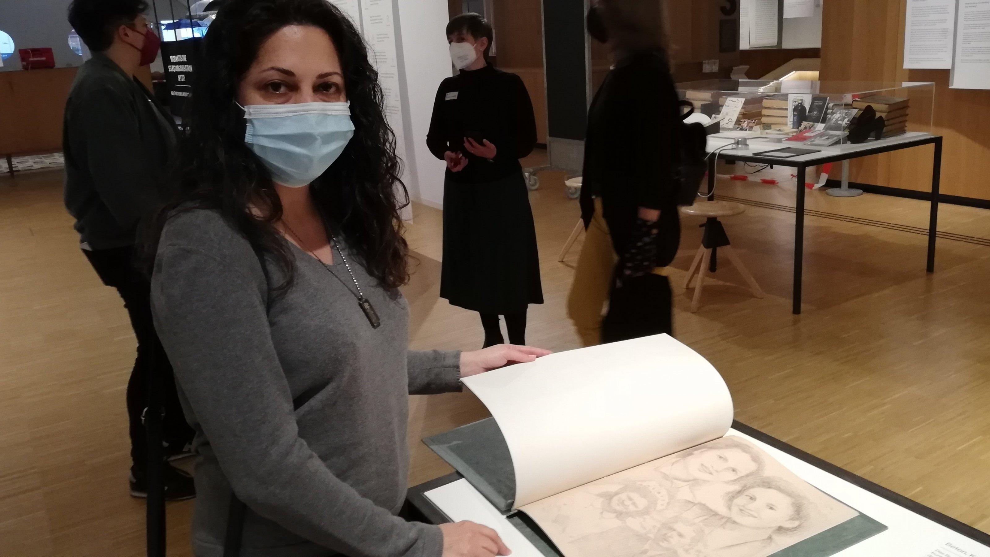 Eine Frau mit langem schwarzen Haar und Corona-Maske blättert in ihrem Buch, das Interviews, Fotomaterial und Zeichnungen enthält.