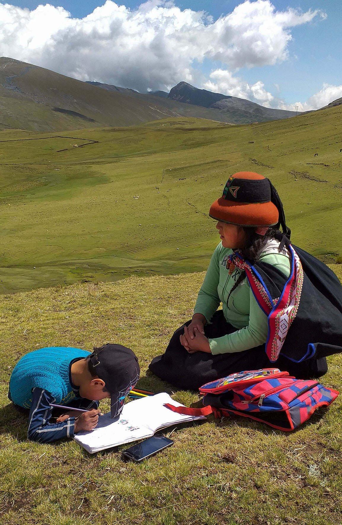 Eine Mutter und ihr Kind sitzen in einer weitern gebirgigen Graslandschaft, das Kind macht Hausaufgaben.