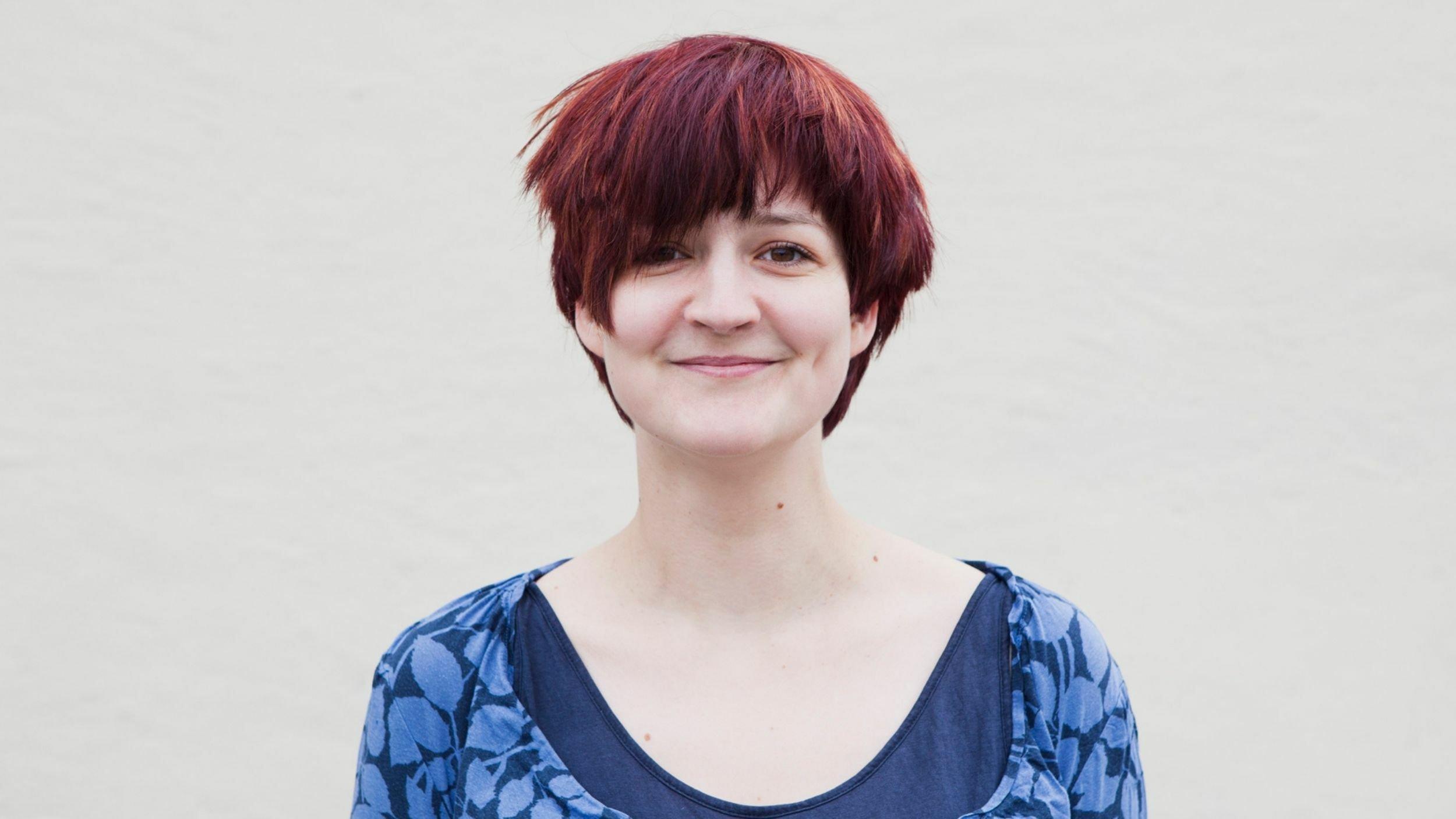 Frau mit rotgefärbten kurzen Haaren vor Wand. Sie trägt ein ausgeschnittenes blau-gemustertes Oberteil.
