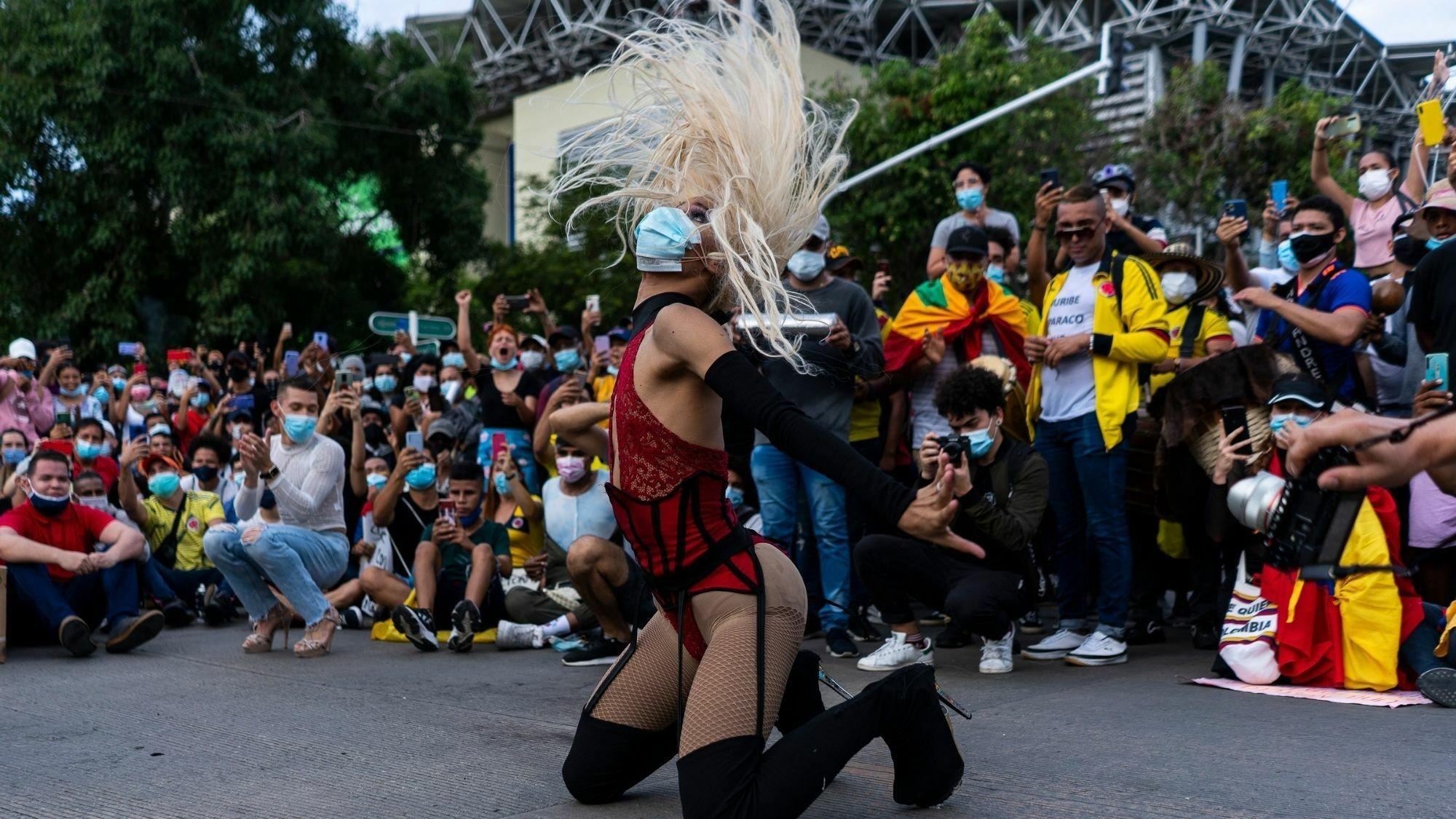 Eine Transfrau mit Mundschutz, hochhackigen Stiefeln und hautengem Spitzenkorsett tanzt auf dem Boden. Sie ist umgeben von  Zuschauerïnnen. Ihre angen Haare stehen vor lauter Schwung senkrecht in der Luft.