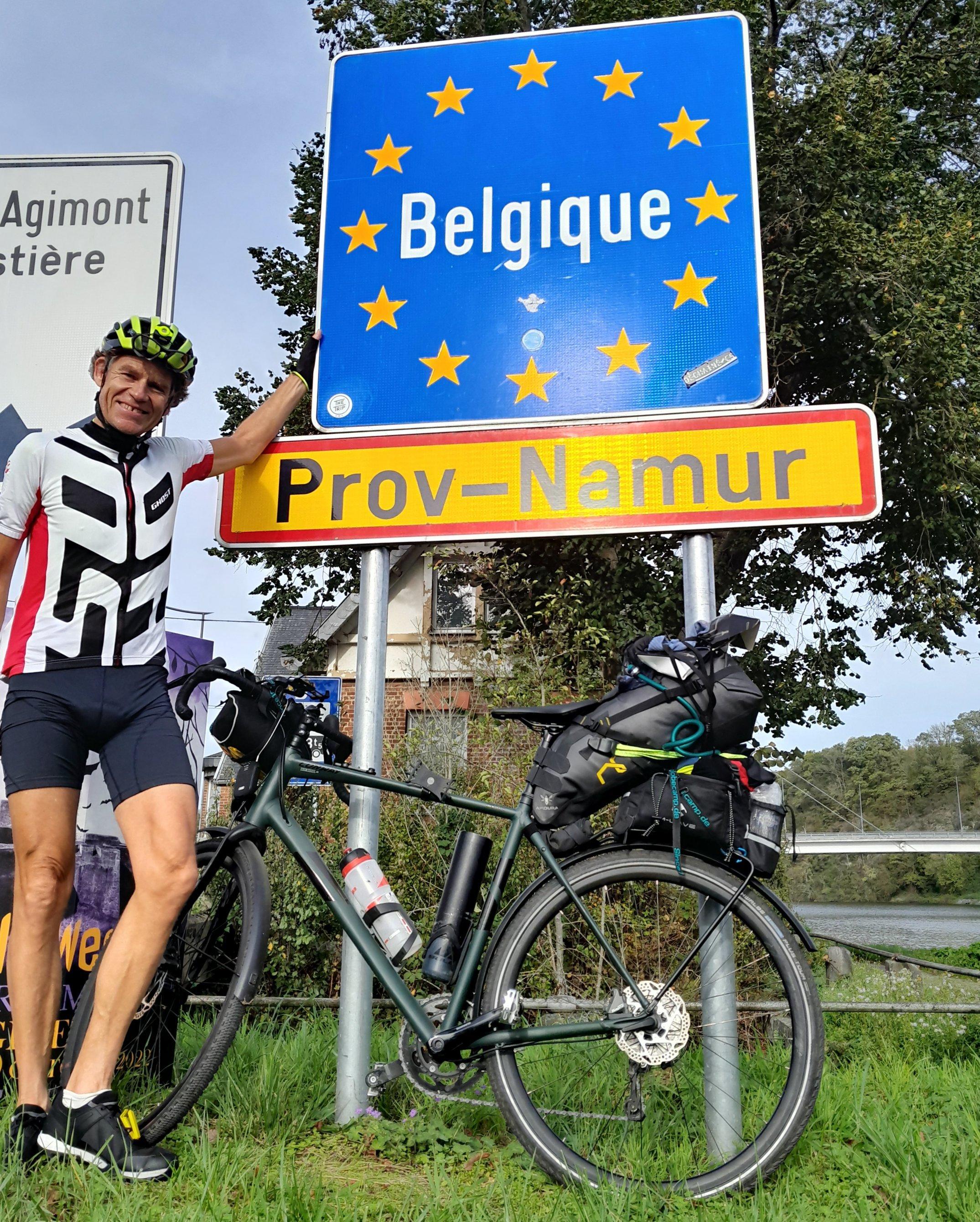 Der Reporter neben dem mit glodenen "Europa-Sternchen" verzierten, blauen Grenzschild, das die weiße Aufschrift "Belgique" trägt.