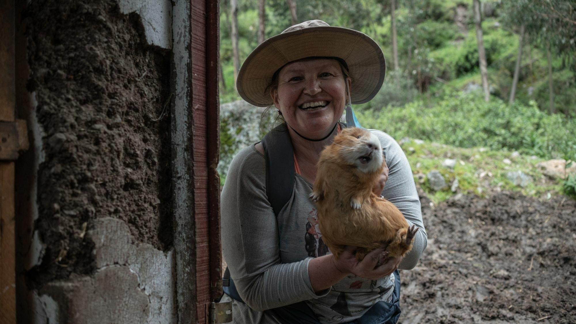 Frau steht im Eingang eines Bauernhauses und hält lachend ein riesiges Meerschwein in die Kamera, das mindestens so groß wie ihr Kopf ist und Zähne zeigt. Sie trägt einen Stoffhut gegen die Höhensonne und sportliche Kleidung.