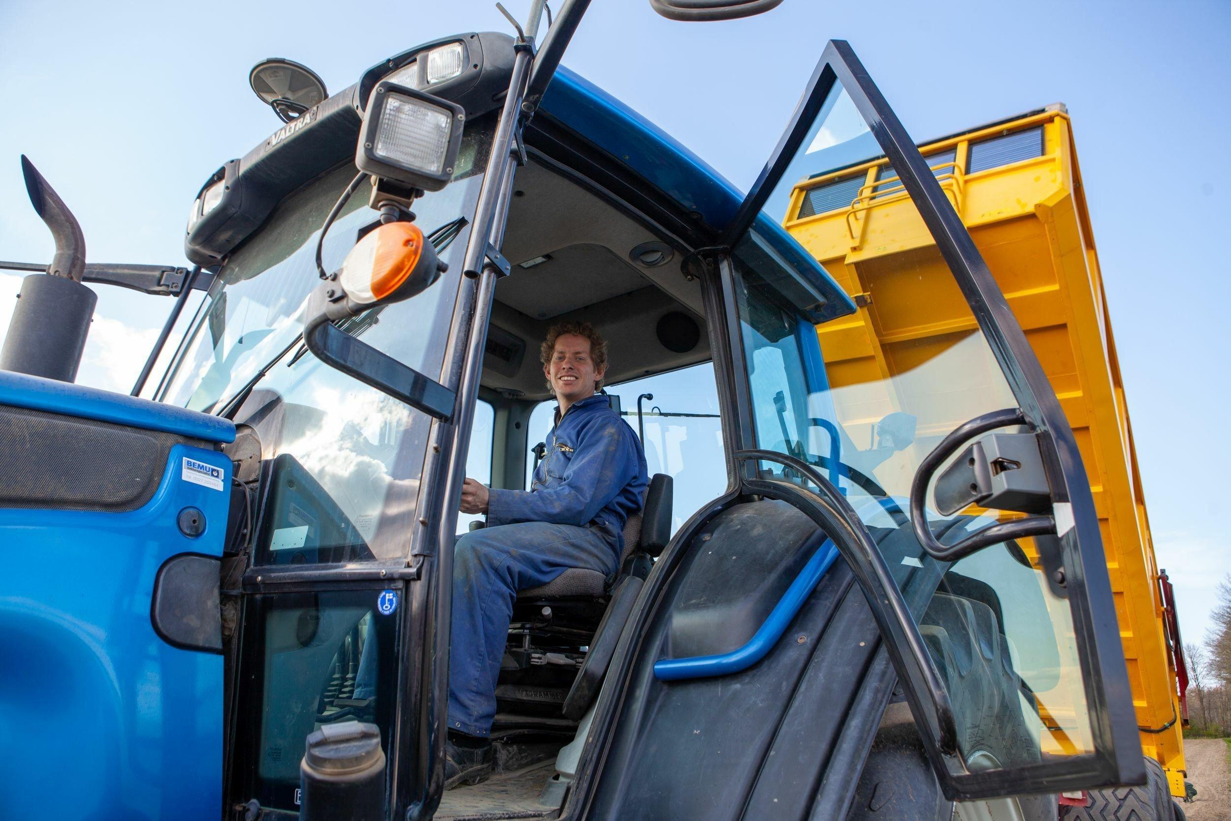 Ein grosser blauer Traktor mit einem gelben Kipplader. Auf dem Traktor ein rund 30-jähriger weisser Mann mit blonden Locken im blauen Overall.