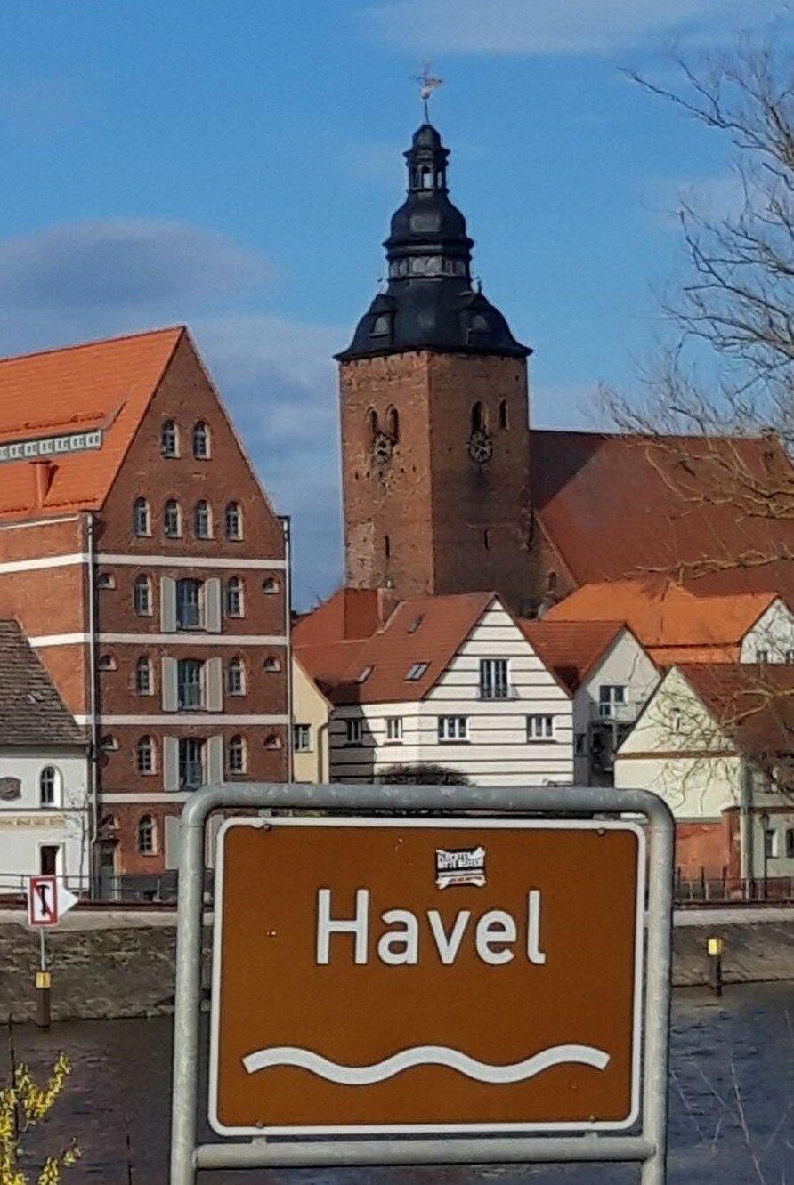 Im Vordergrund ist die Havel und ein entsprechendes Schild mit Wellensymbol zu sehen, im Hintergrund Altstadthäuser, unter anderem ein hanseatisches Kaufmannshaus, sowie eine Kirche.