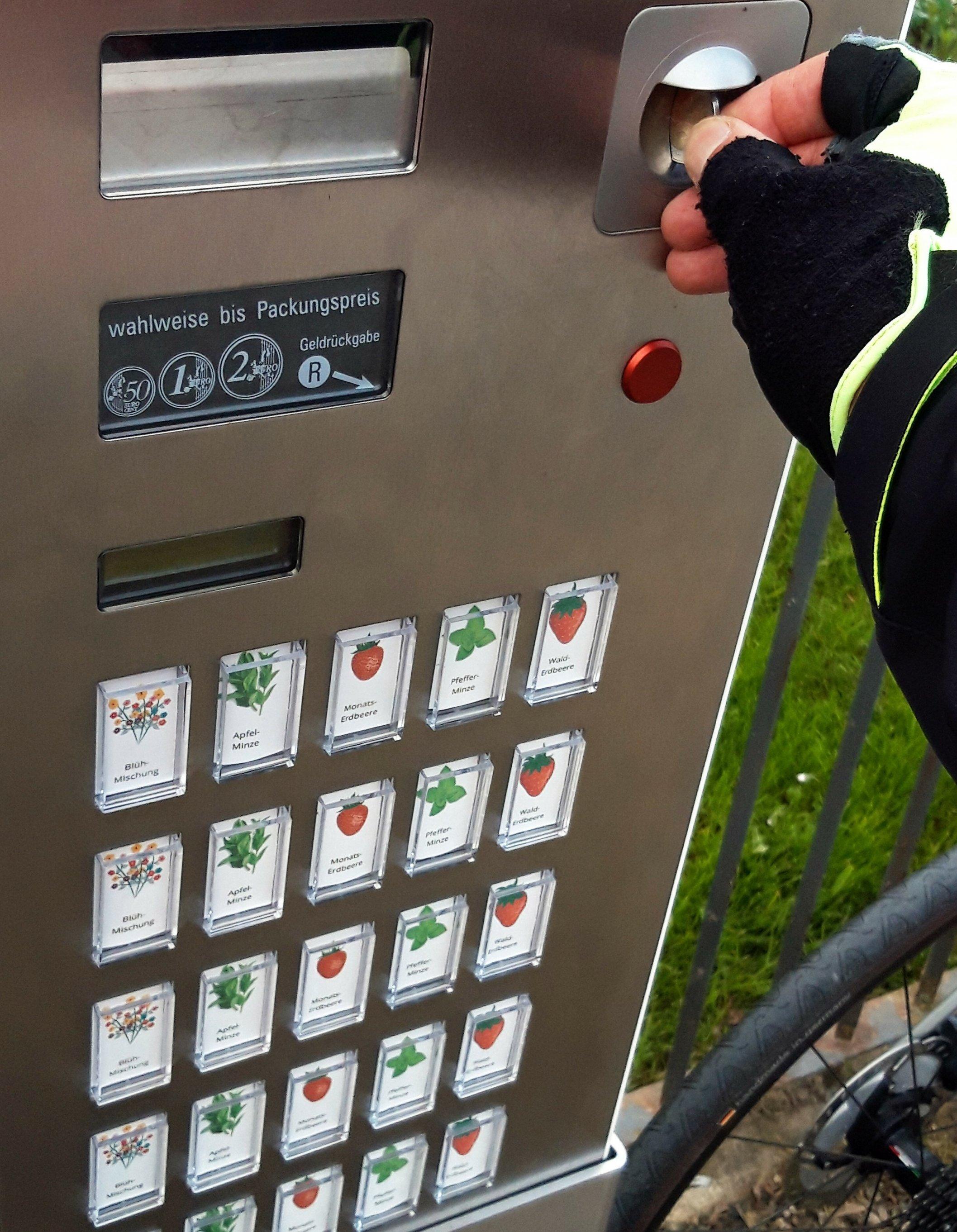 Eine Hand schiebt eine 2-Euro-Münze in den Automatenschlitz, unter dem farbenfroh die Wahltasten für verschiedene Blumensamen zu sehen sind.