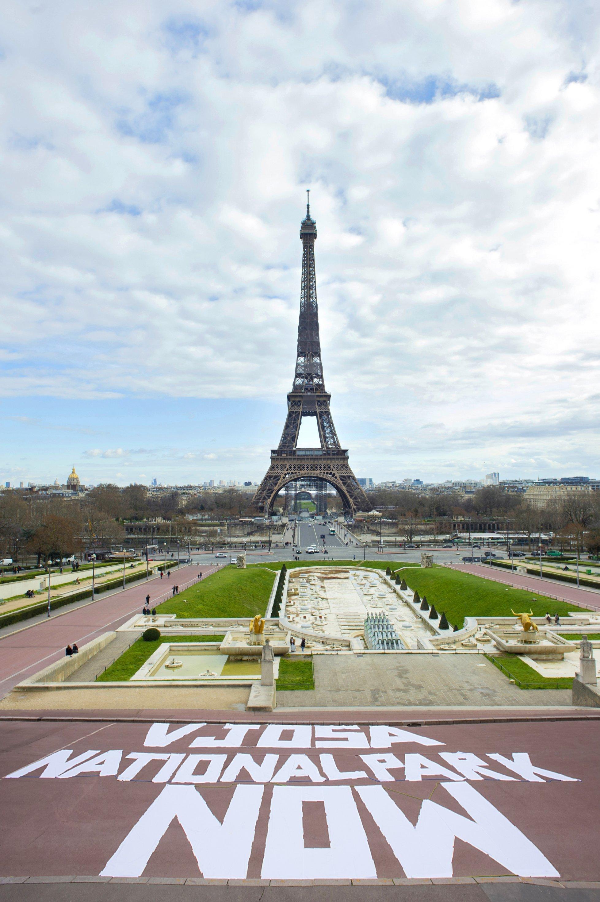 Weißer Schriftzug auf einem roten Platz, dahinter dem Eiffelturm, blauer Himmel mit Wolken