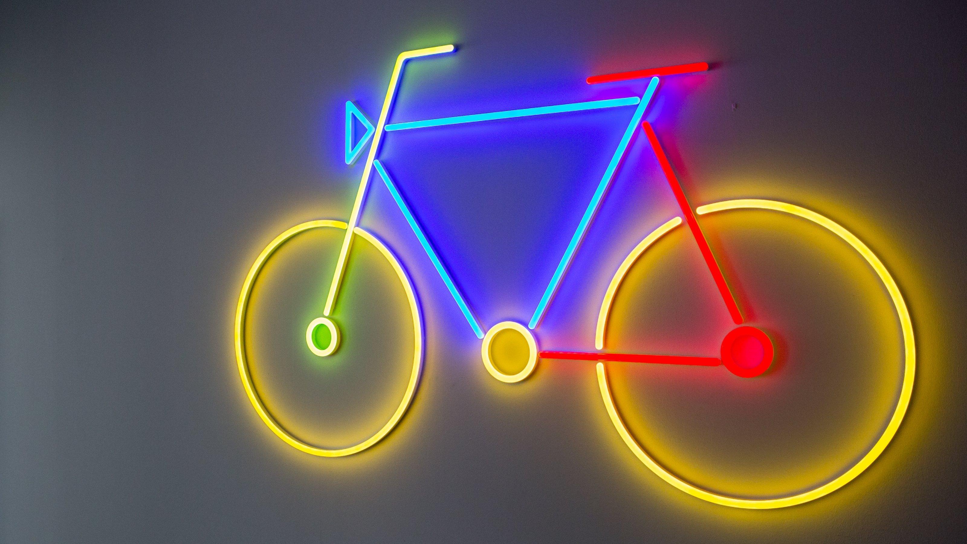 Stilisiertes Fahrrad, das in Neonfarben gelb, rot, blau und grün vor einem dunklen Hintergrund leuchtet.