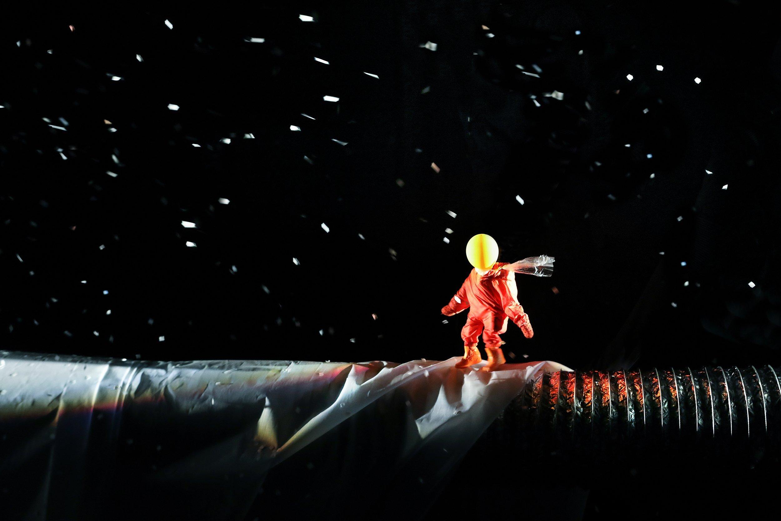 Auf einem großen Lüftungsrohr spaziert eine kleine Figur mit einem gelben Luftballon als Kopf durch ein künstliches Schneegestöber durch die Nacht. Die Puppenspieler:innen im Hintergrund sieht man nicht.
