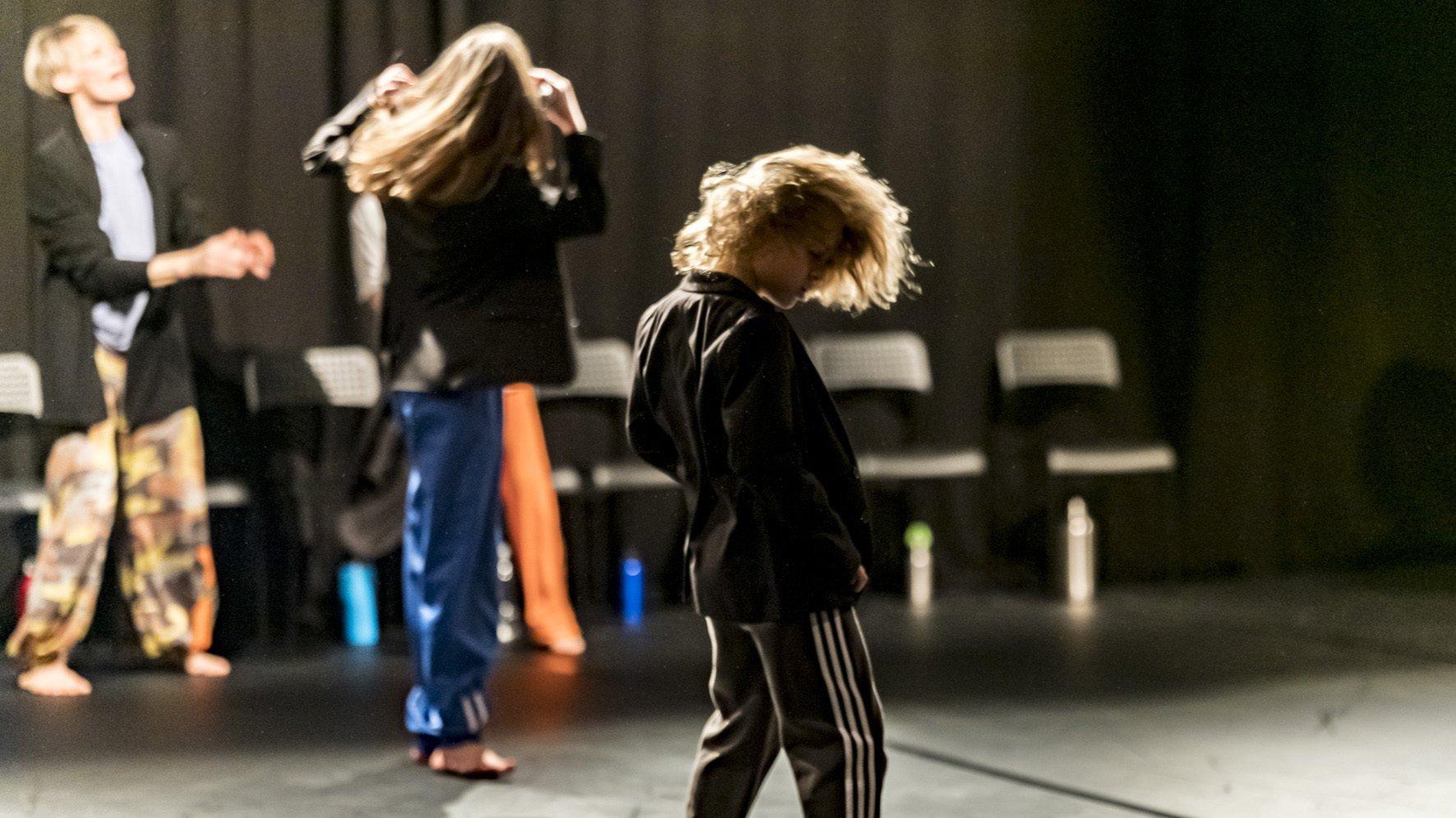 Auf der Bühne sieht man einen kleinen Jungen wild tanzen. Im Hintergrund bewegen sich noch zwei weitere Menschen, ein sehr junger so schnell, dass die Konturen verwischen. Es ist eine Szene aus dem Tanzstück „Mischpoke“ von Barbara Fuchs.