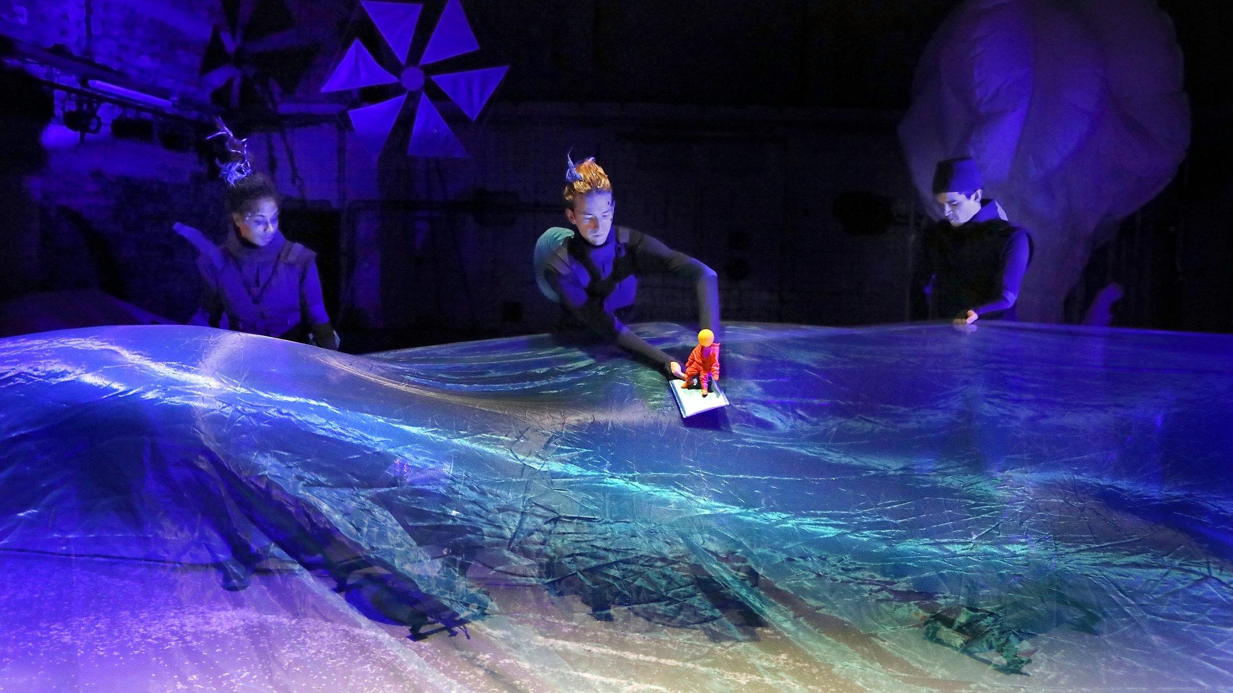 Abenaa Prempeh, Markus Schabbing und William Cooper performen „Es liegt was in der Luft“ auf der Bühne: Eine kleine Figur schwimmt über einen Ozean aus einer Folie.