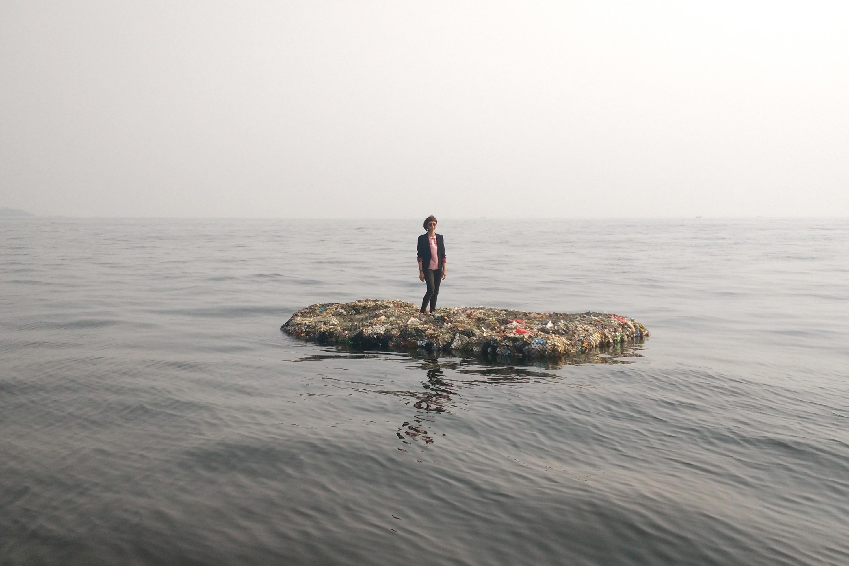 Ein Floß aus Plastikmüll treibt auf ruhiger See. Darauf steht die Künstlerin Tita Salina.